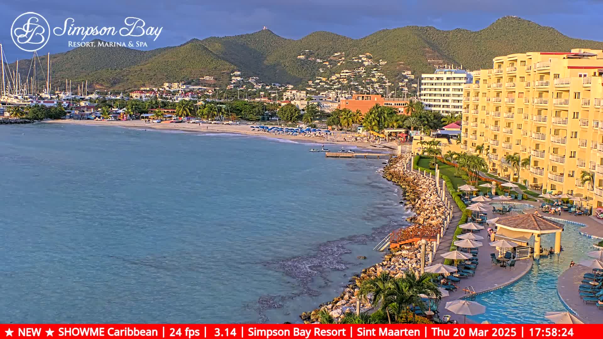 A tranquil beach scene with calm, turquoise water, a sandy shore lined with beach umbrellas, a resort with a pool, and a mountainous backdrop under a mostly clear sky.
