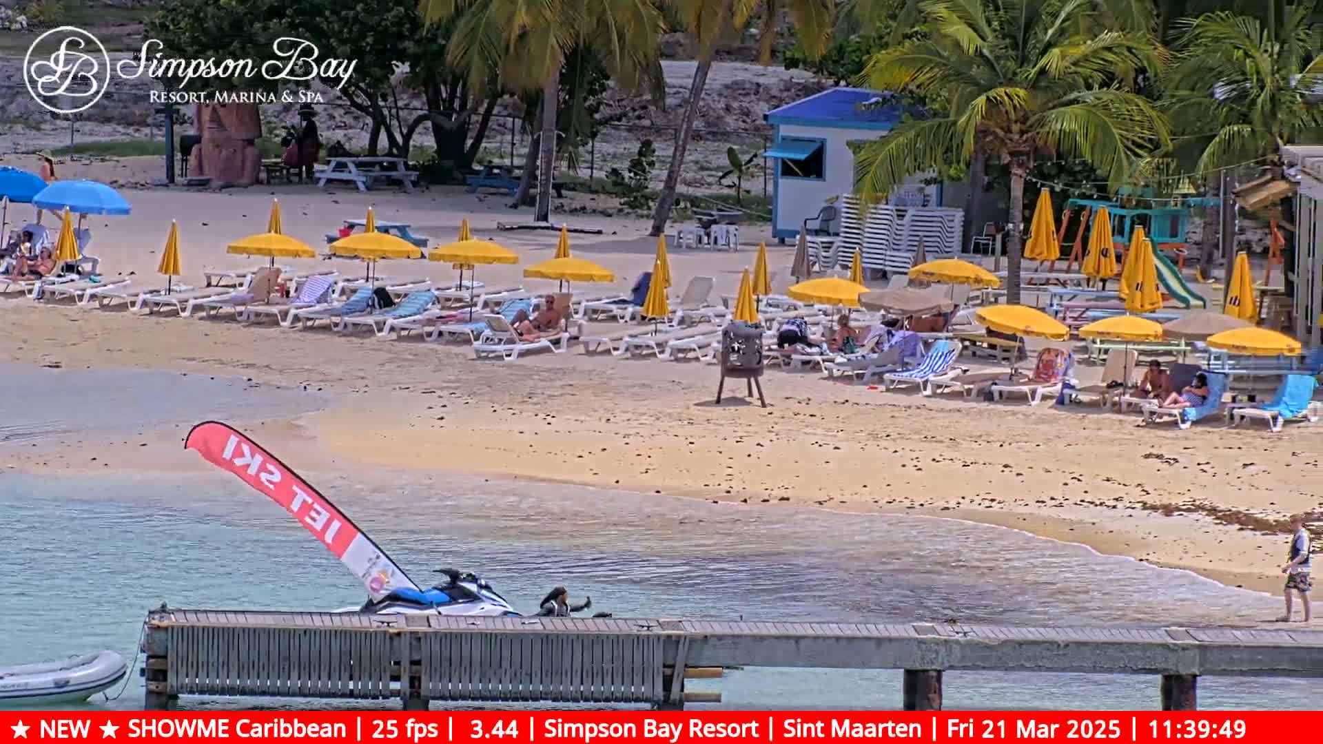 A sandy beach with numerous yellow umbrellas and lounge chairs, a jet ski near a dock, and calm, shallow water under a sunny sky.