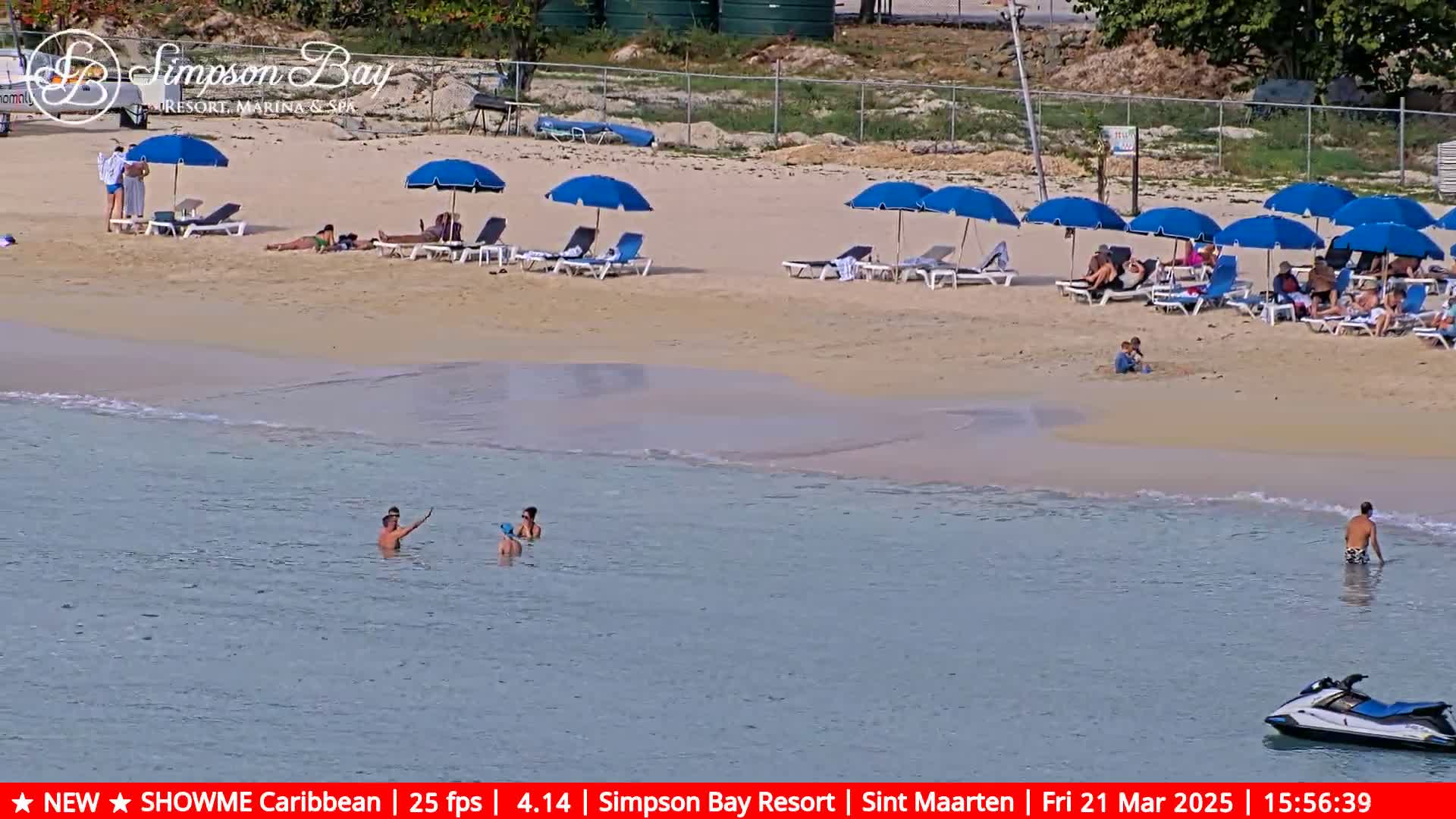 A sandy beach with calm, clear water is populated by bathers and sunbathers under blue beach umbrellas.