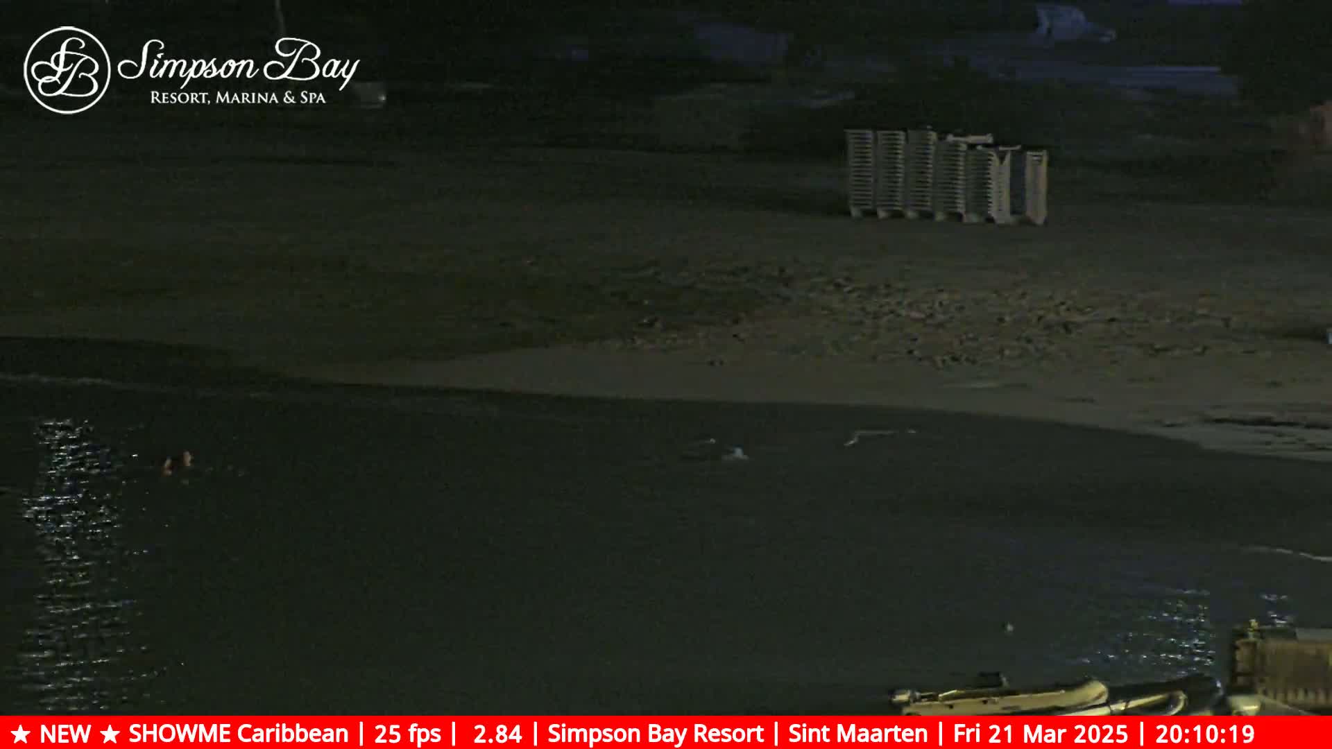 At night, two people swim in dark water near a sandy beach with some beach chairs stacked up on the shore.