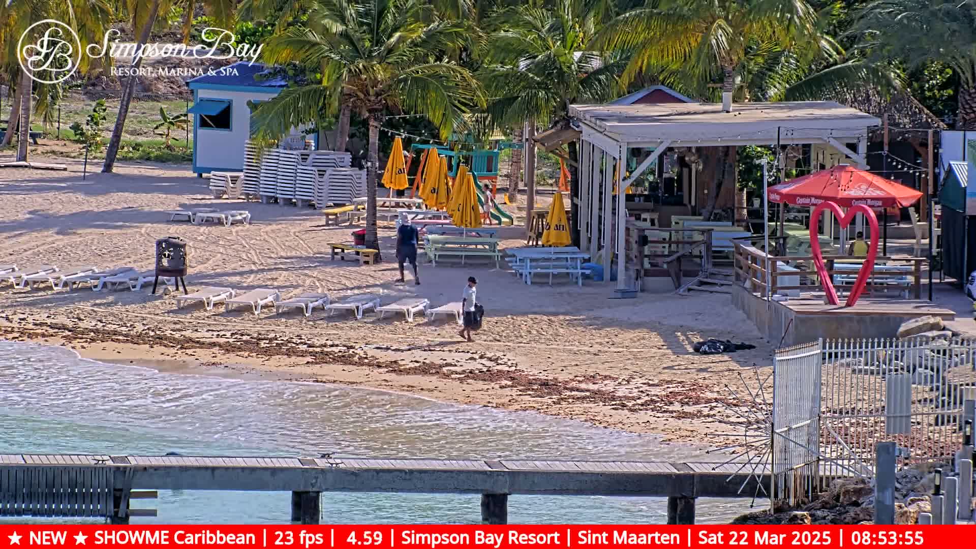 A sandy beach with lounge chairs, a beachfront bar and restaurant, and several people walking, all under a sunny sky.