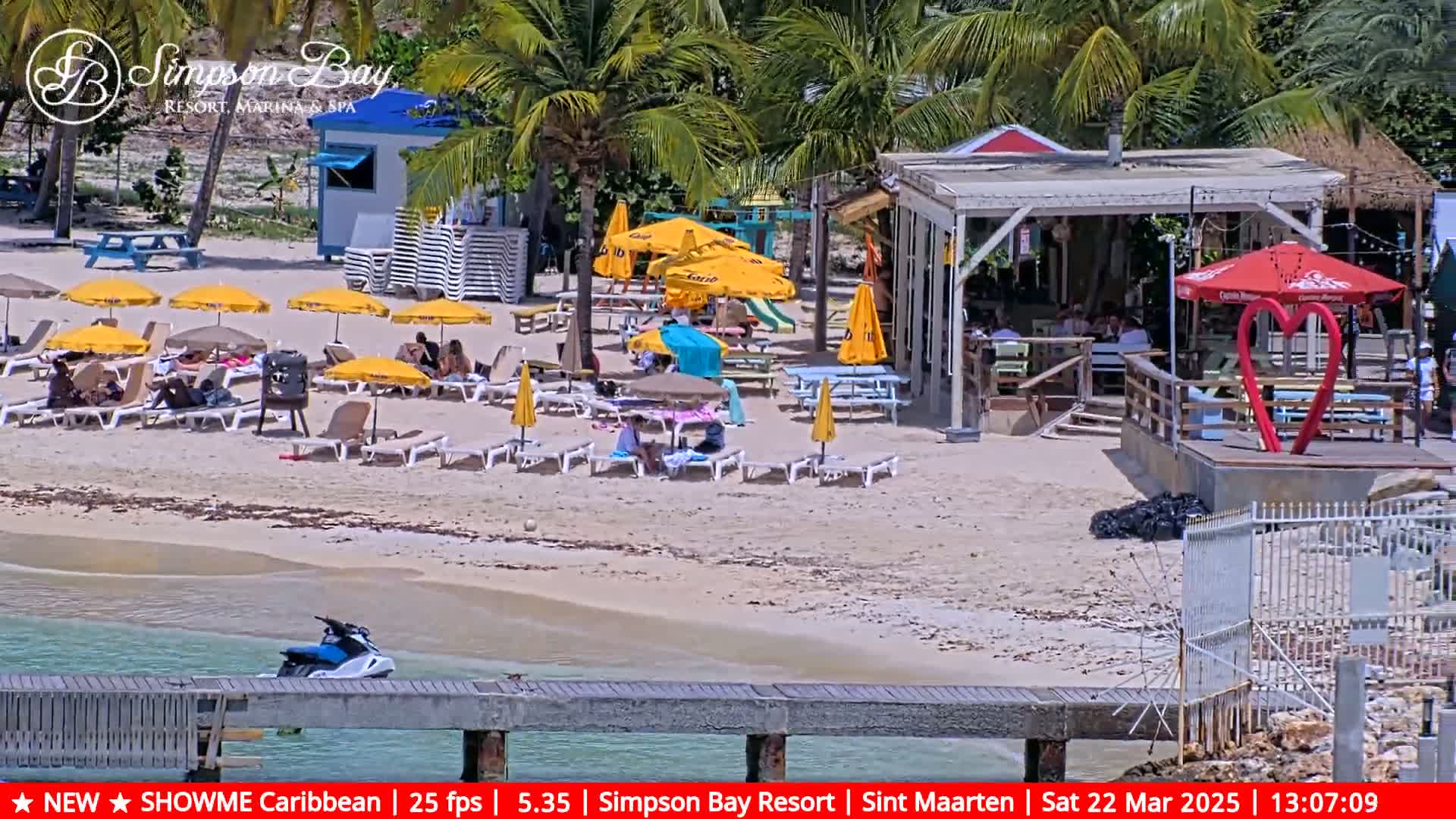 A sunny beach scene shows people relaxing on lounge chairs under yellow umbrellas near a beach bar and a jet ski docked at a pier.
