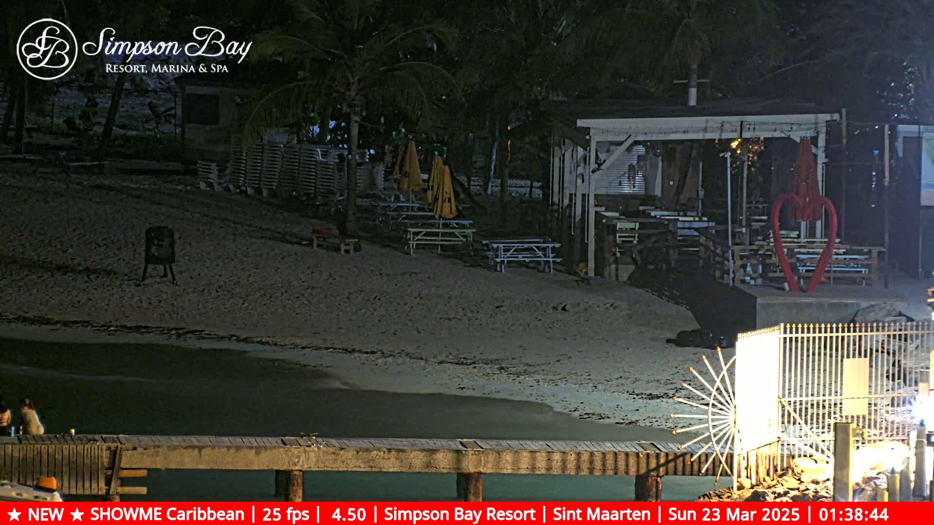 A nighttime beach scene shows a sandy shoreline, a partially visible building with picnic tables and umbrellas, and a wooden pier, all under dark conditions.