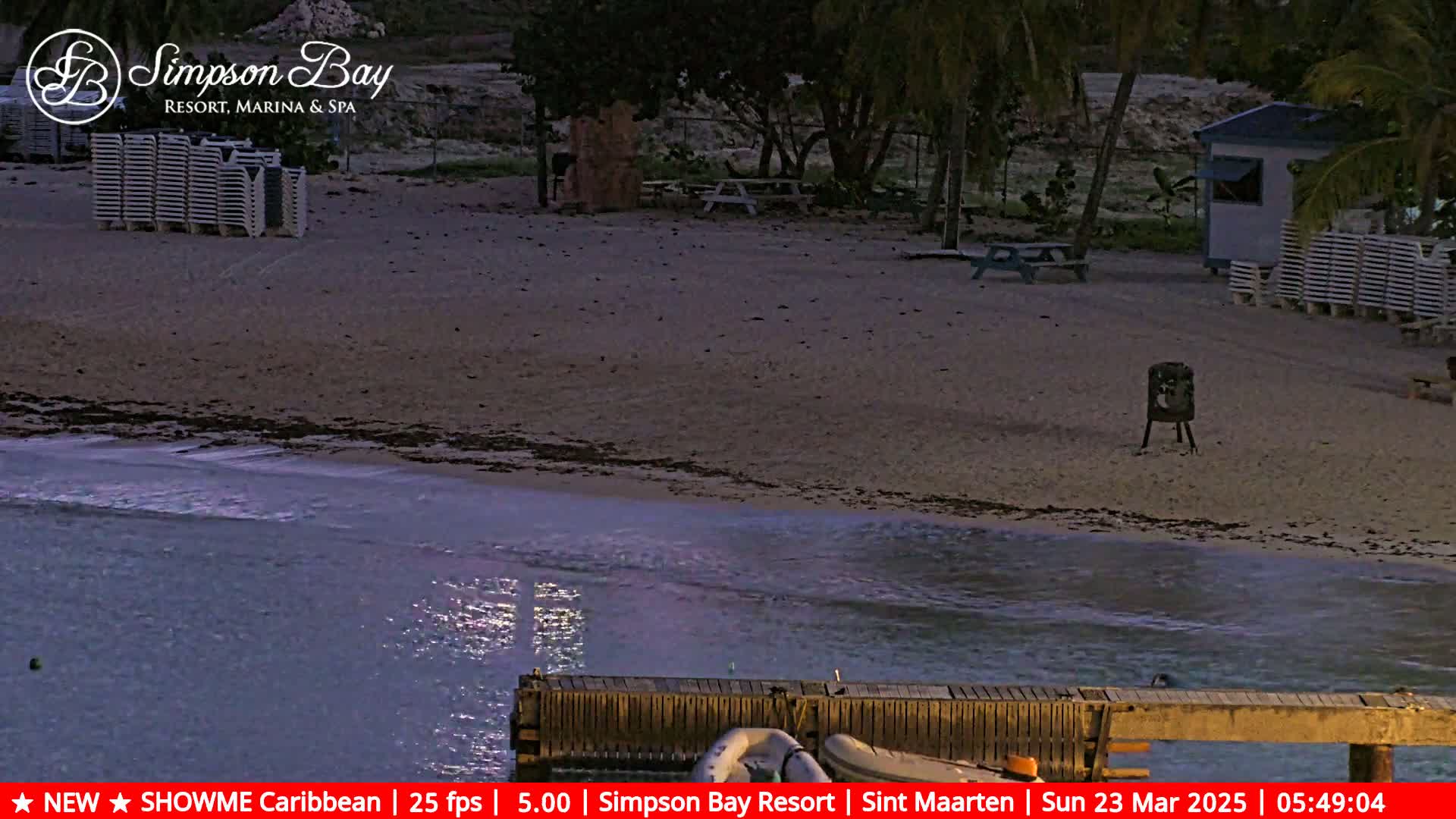 A sandy beach at dusk, with calm water lapping the shore, features stacked lounge chairs, a small building, and picnic tables.