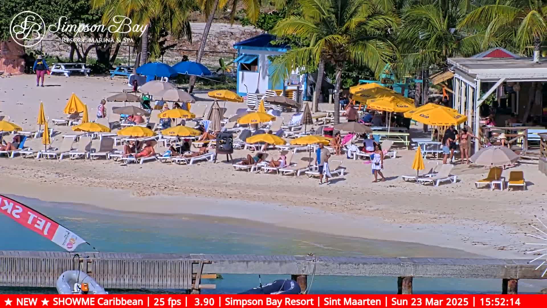 A sunny beach scene shows numerous beach umbrellas, lounge chairs, and people relaxing on a sandy beach near a small building and a pier extending into calm, clear water.