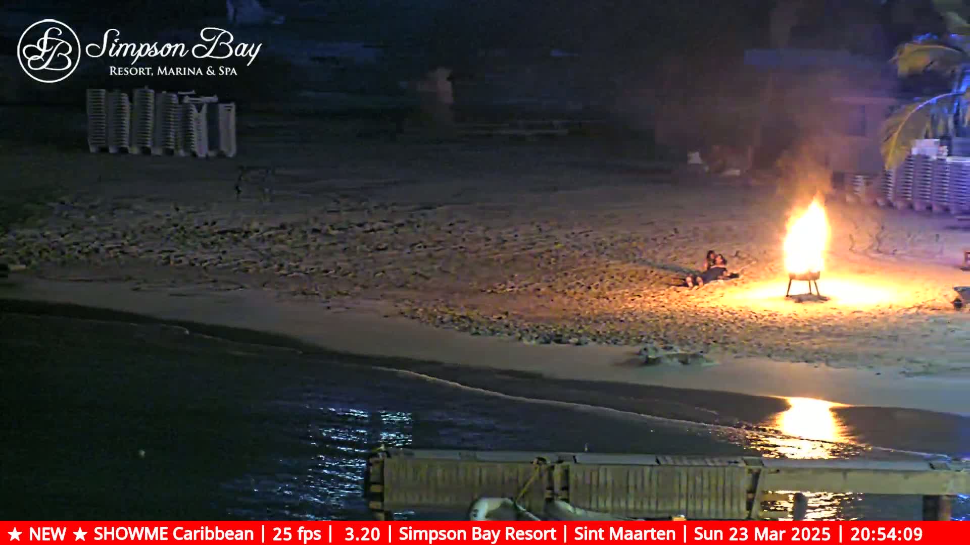 A bonfire burns brightly on a sandy beach at night, next to the ocean, with a couple sitting nearby.