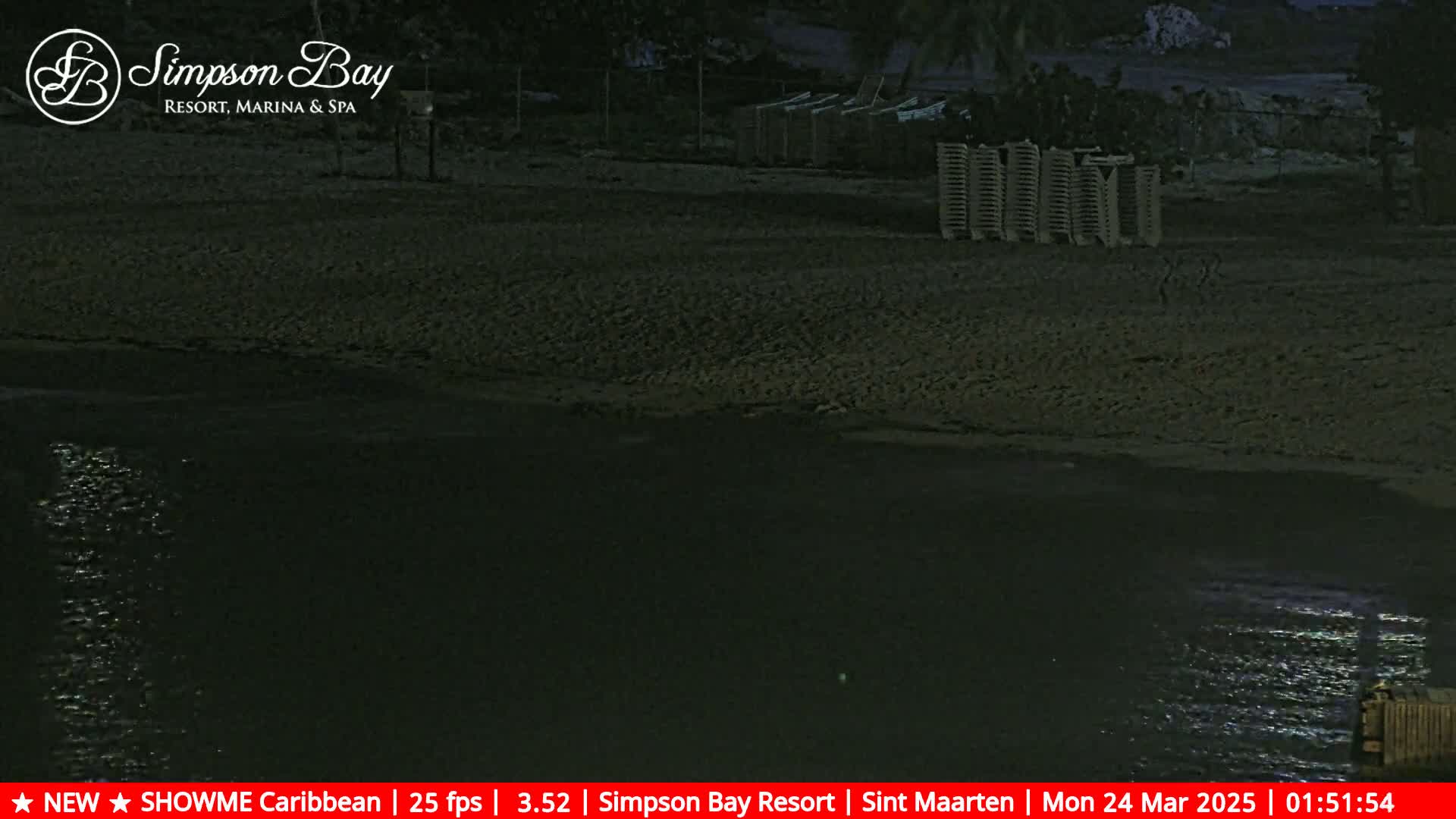 A dark, nighttime scene shows a sandy beach with some beach chairs, next to dark water reflecting light.