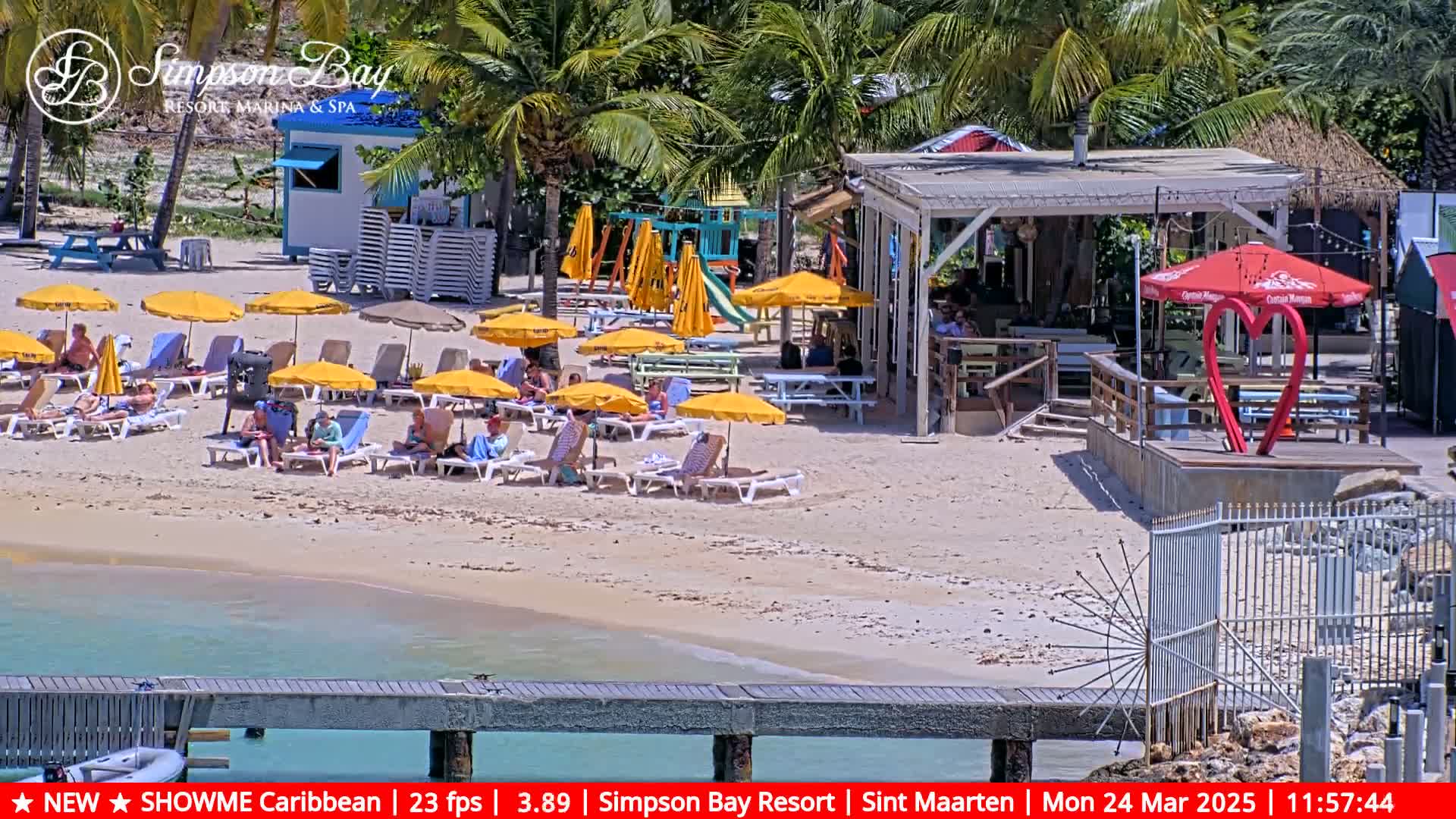 A sunny beach scene shows people relaxing under yellow umbrellas on lounge chairs near a beachfront bar and playground.