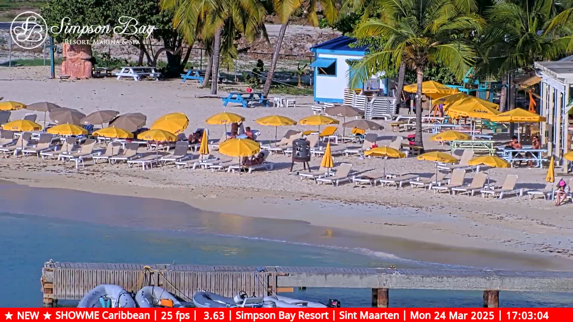 A sunny beach scene shows numerous yellow umbrellas and lounge chairs set up on a sandy beach near a small building and a wooden dock.