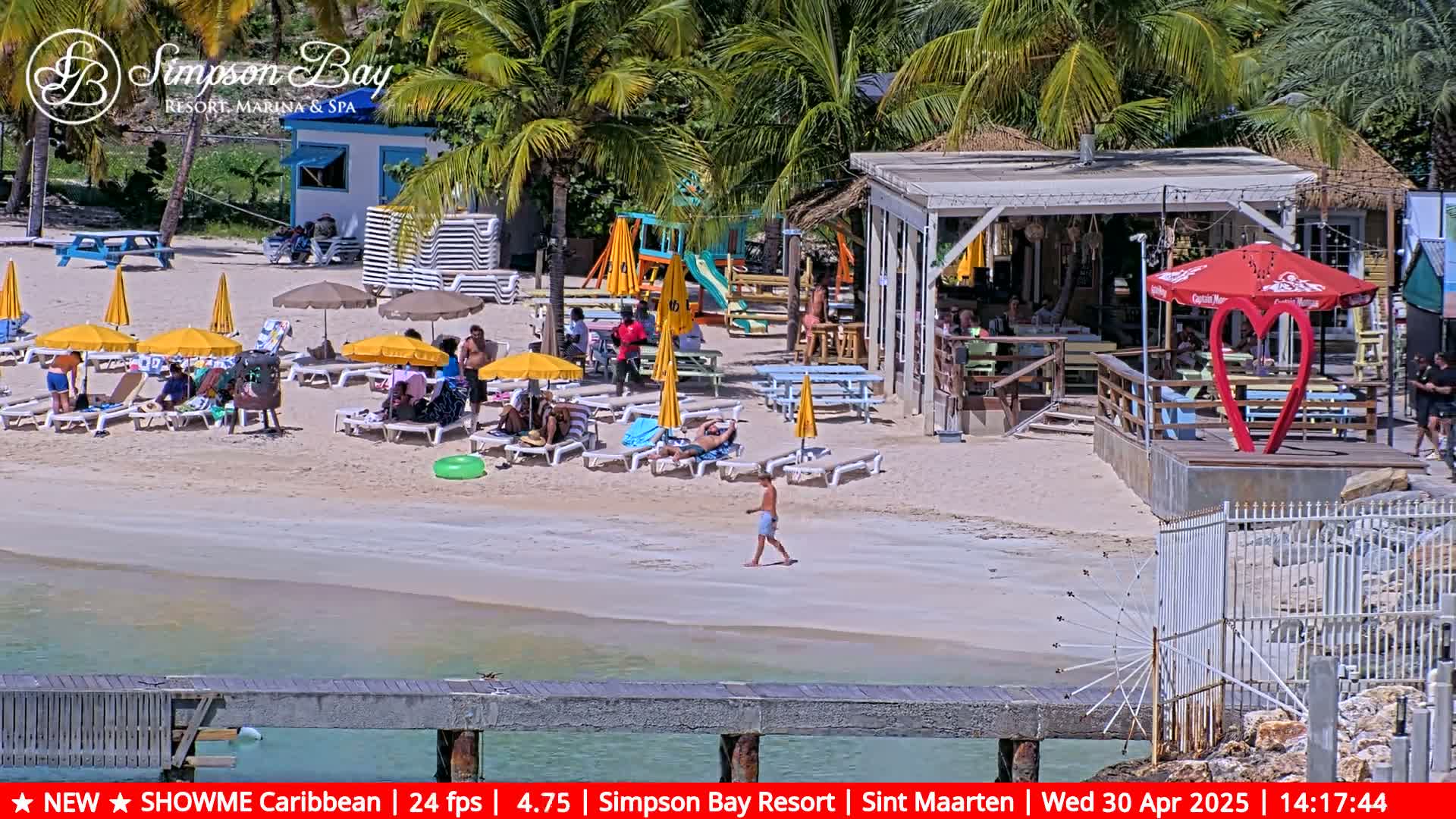 A sandy beach scene shows people relaxing under yellow umbrellas near a beachfront bar and playground on a sunny day.