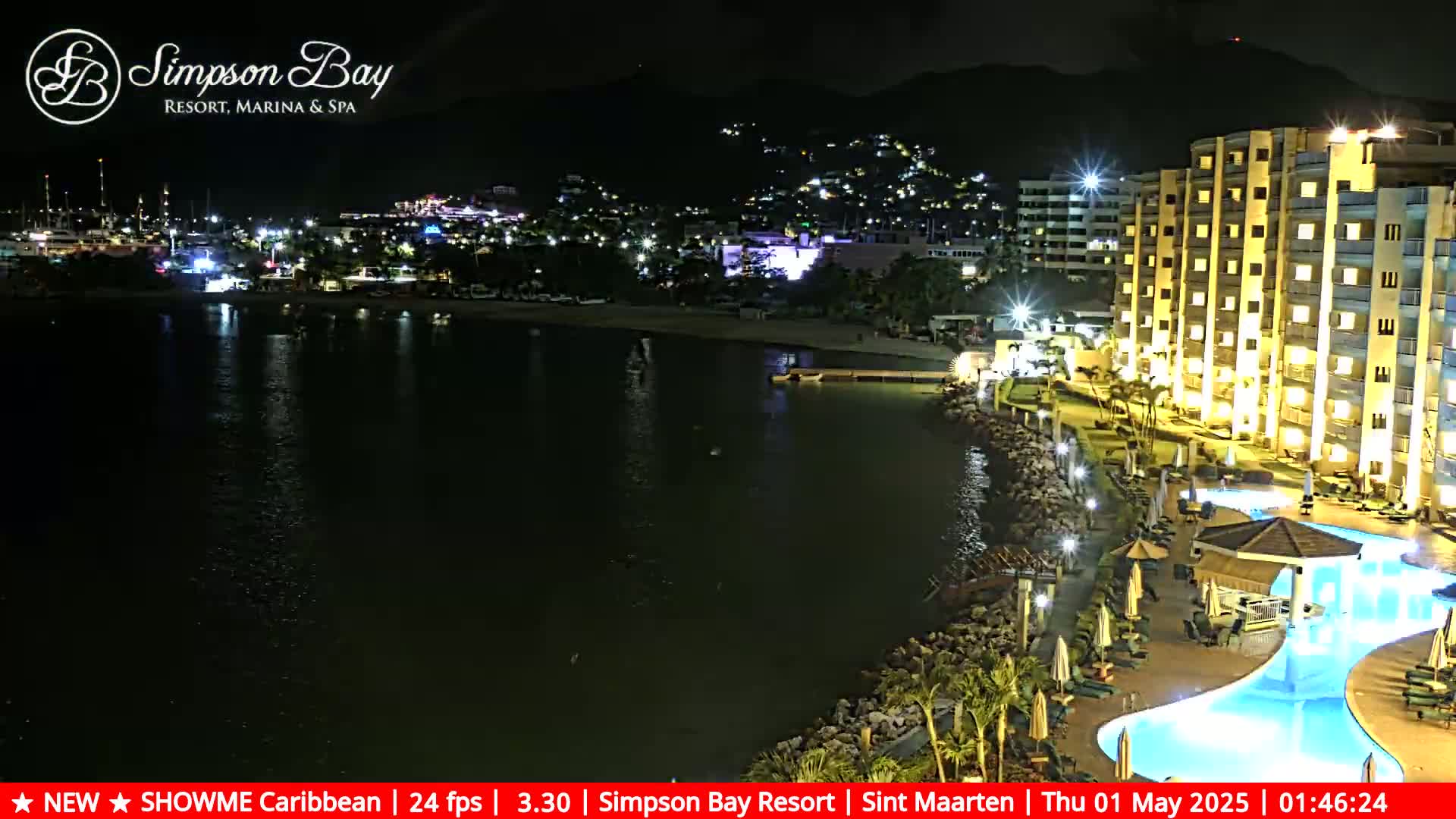 A brightly lit resort with a swimming pool sits on the waterfront at night, overlooking a dark body of water and a town illuminated with lights.