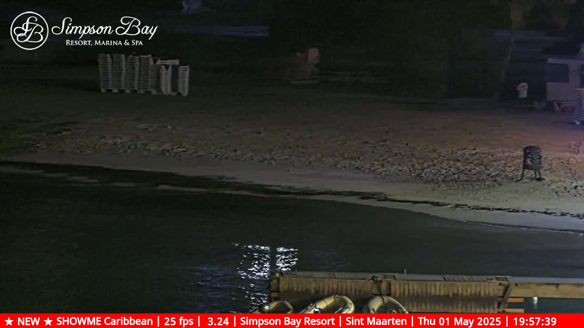 A dark, nighttime beach scene shows calm water lapping at a sandy shore, with some beach chairs stacked in the background.