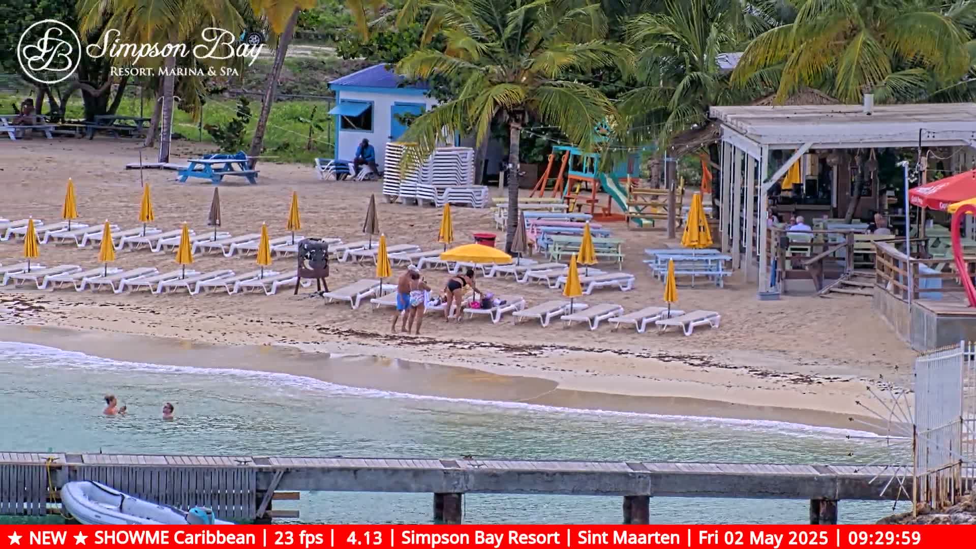A sandy beach with lounge chairs and umbrellas, a small beach bar, and a playground is shown next to calm, clear water.