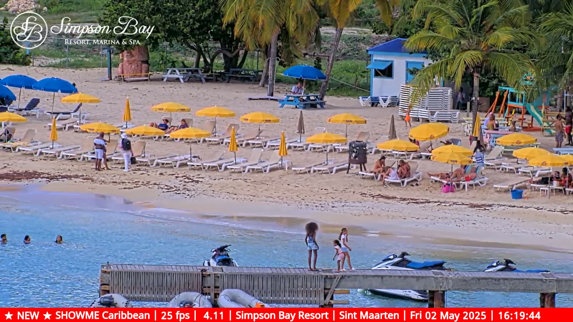 A sandy beach with numerous yellow umbrellas and lounge chairs, people relaxing and swimming in the calm, clear water, and jet skis docked at a pier.