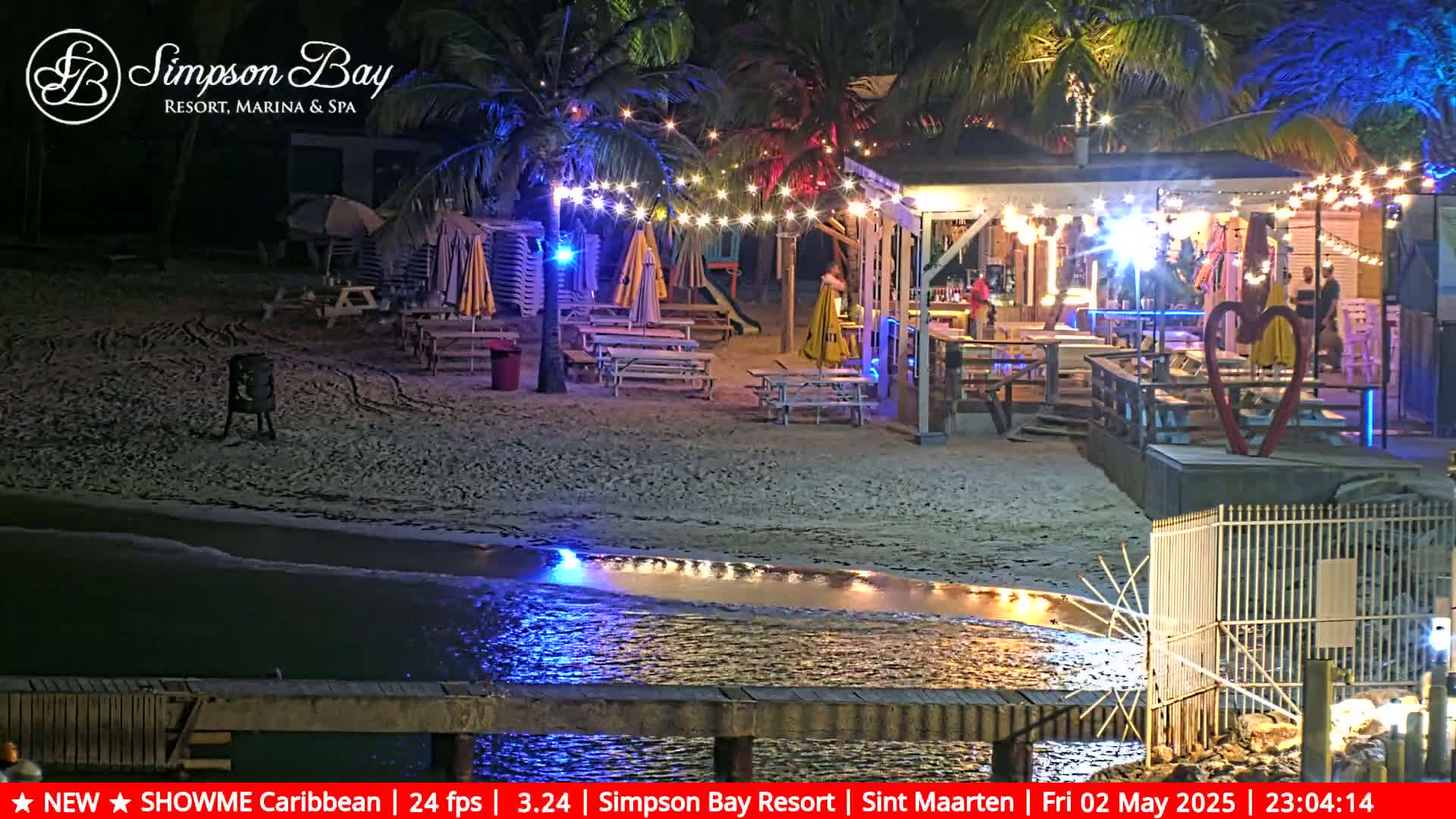 A beachside bar and restaurant at night, illuminated by string lights, is situated on a sandy beach next to calm, shallow water.