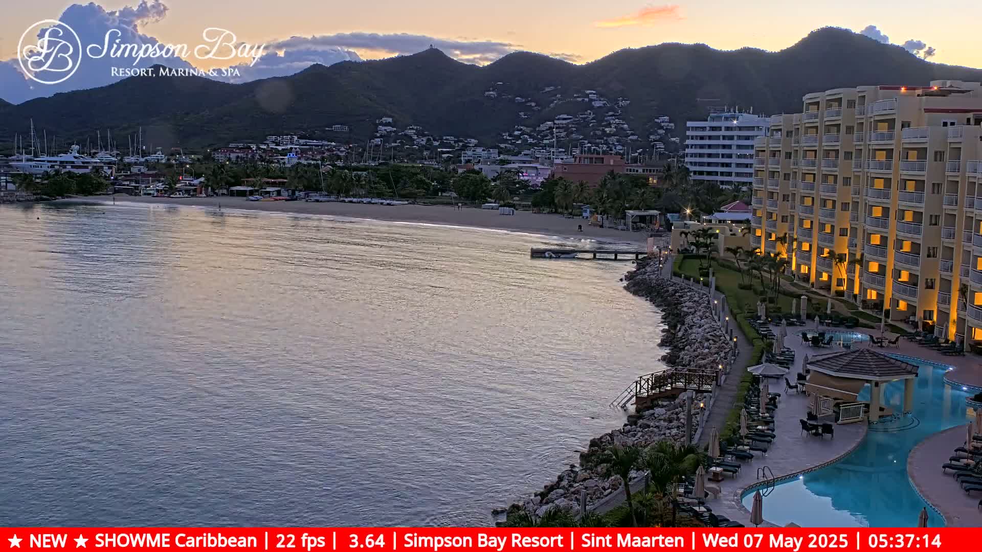 A calm bay at sunset, with a sandy beach, a resort with a lit-up pool area, and a mountainous backdrop.