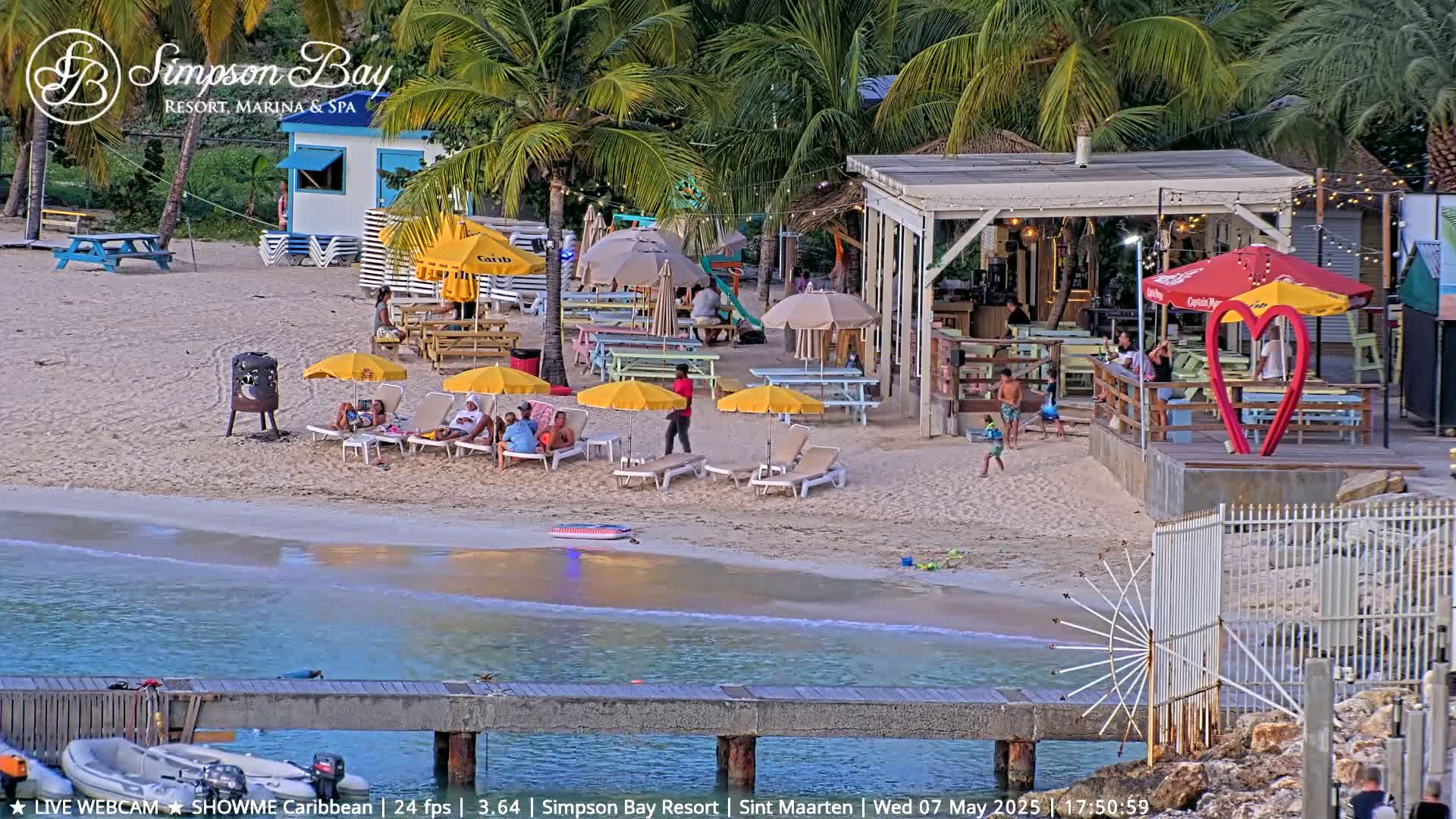 A beach scene at sunset shows people relaxing under yellow umbrellas on a sandy beach near a bar and restaurant with a pier extending into calm turquoise water.