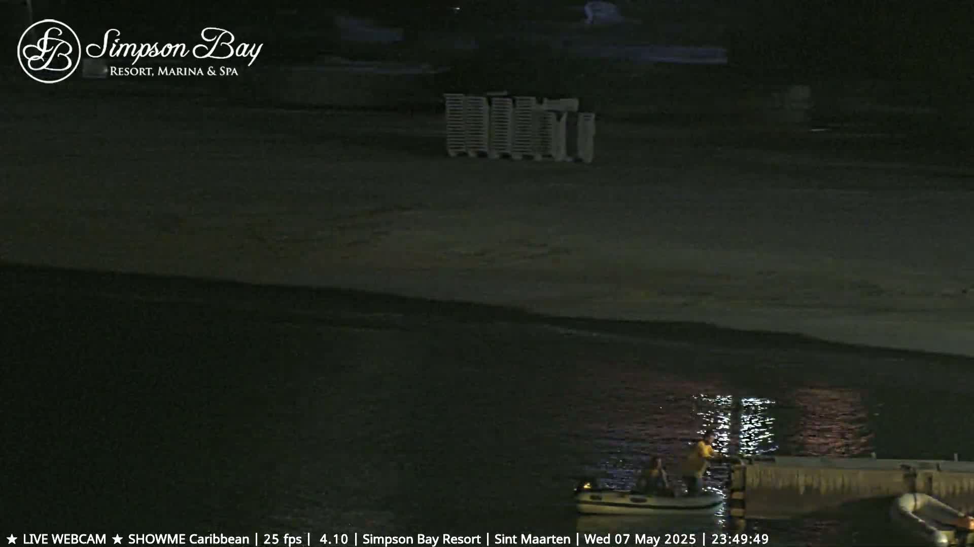 At night, two people are on a small inflatable boat at a dock near a dark sandy beach with folded chairs.