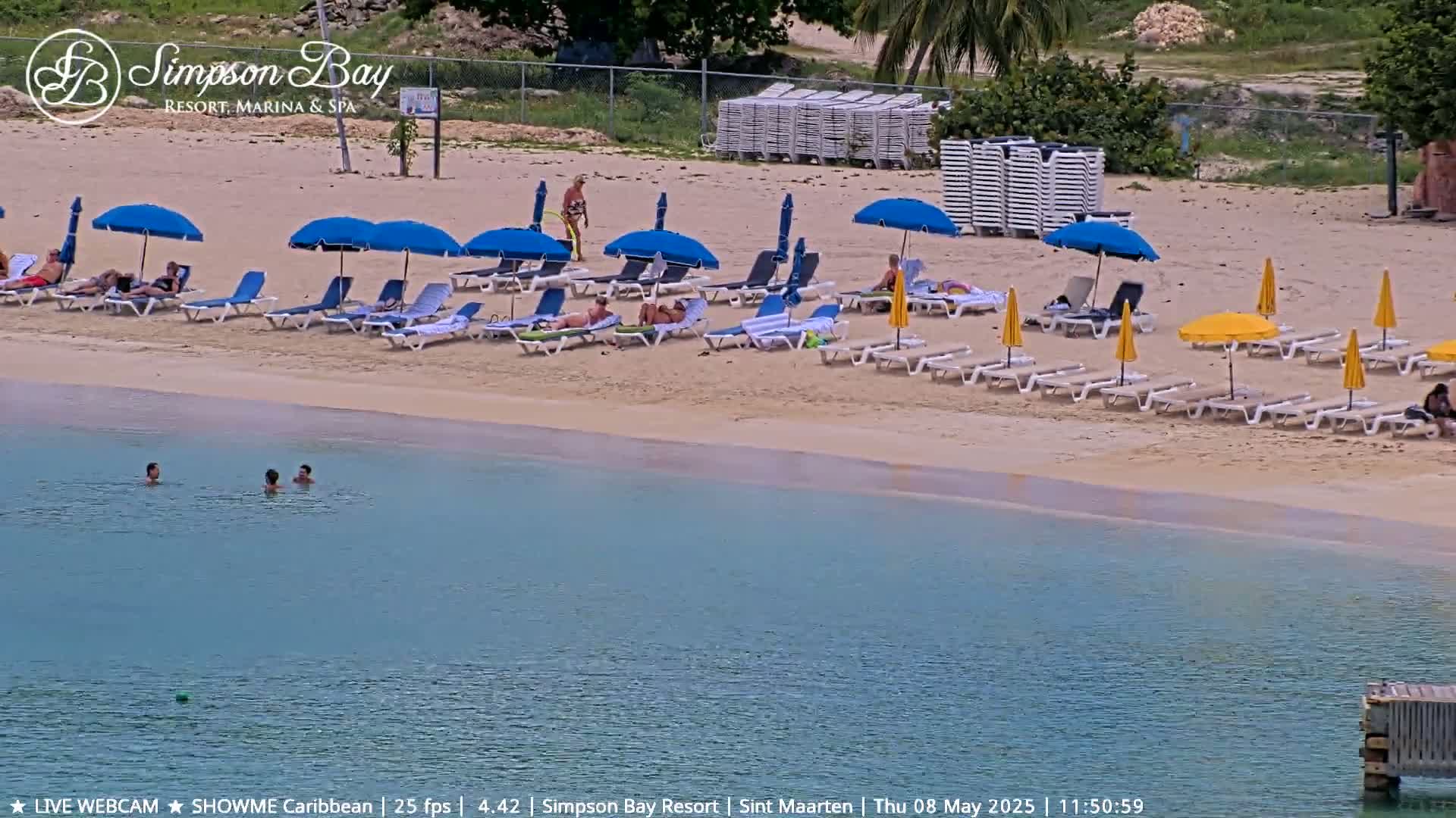 A sandy beach with numerous blue and yellow umbrellas, lounge chairs, and people relaxing, a calm turquoise body of water in front of it, and several people swimming in the shallows.