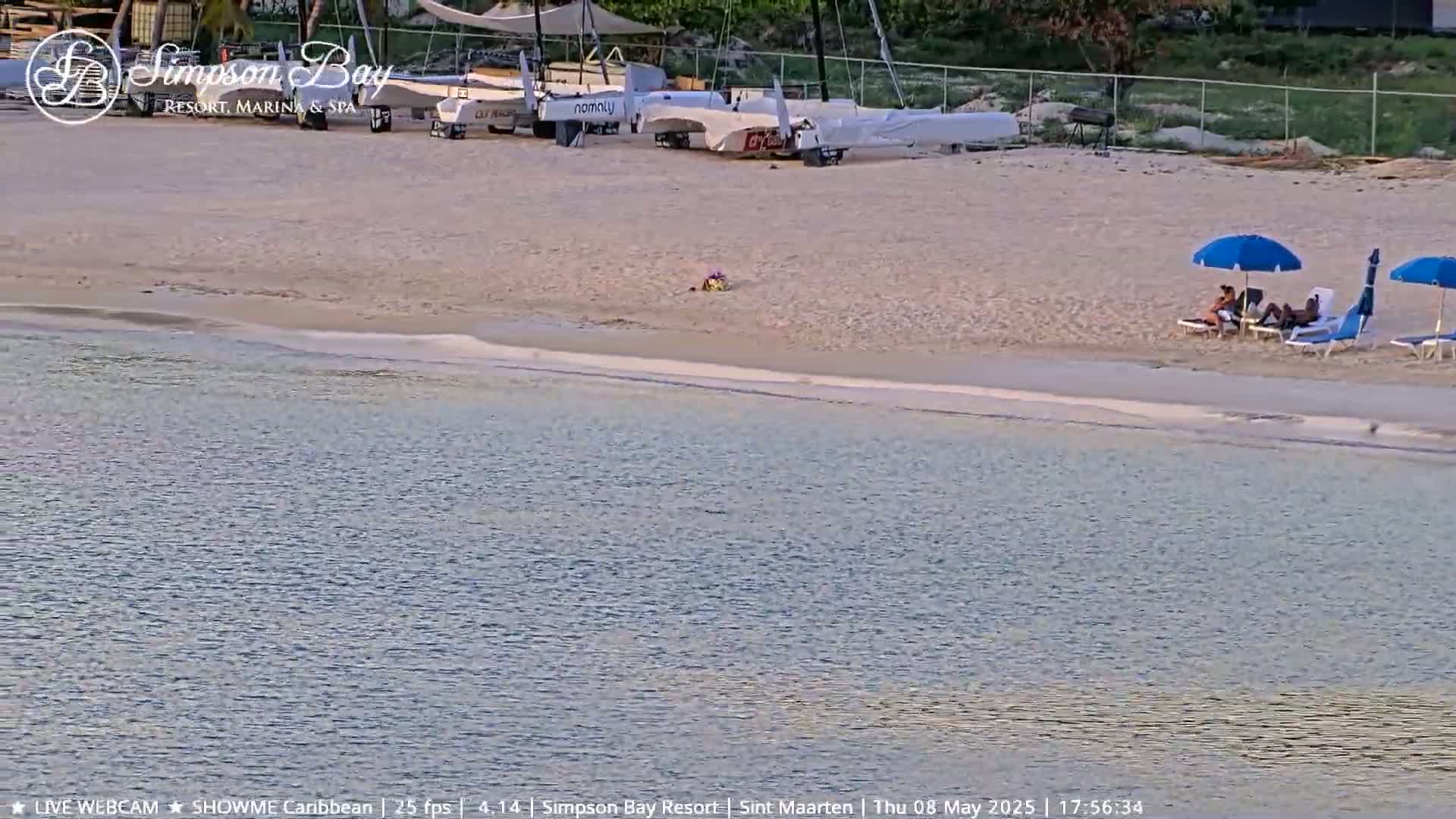 A sandy beach with several sailboats on the shore, two people relaxing under umbrellas, and calm, shallow water.