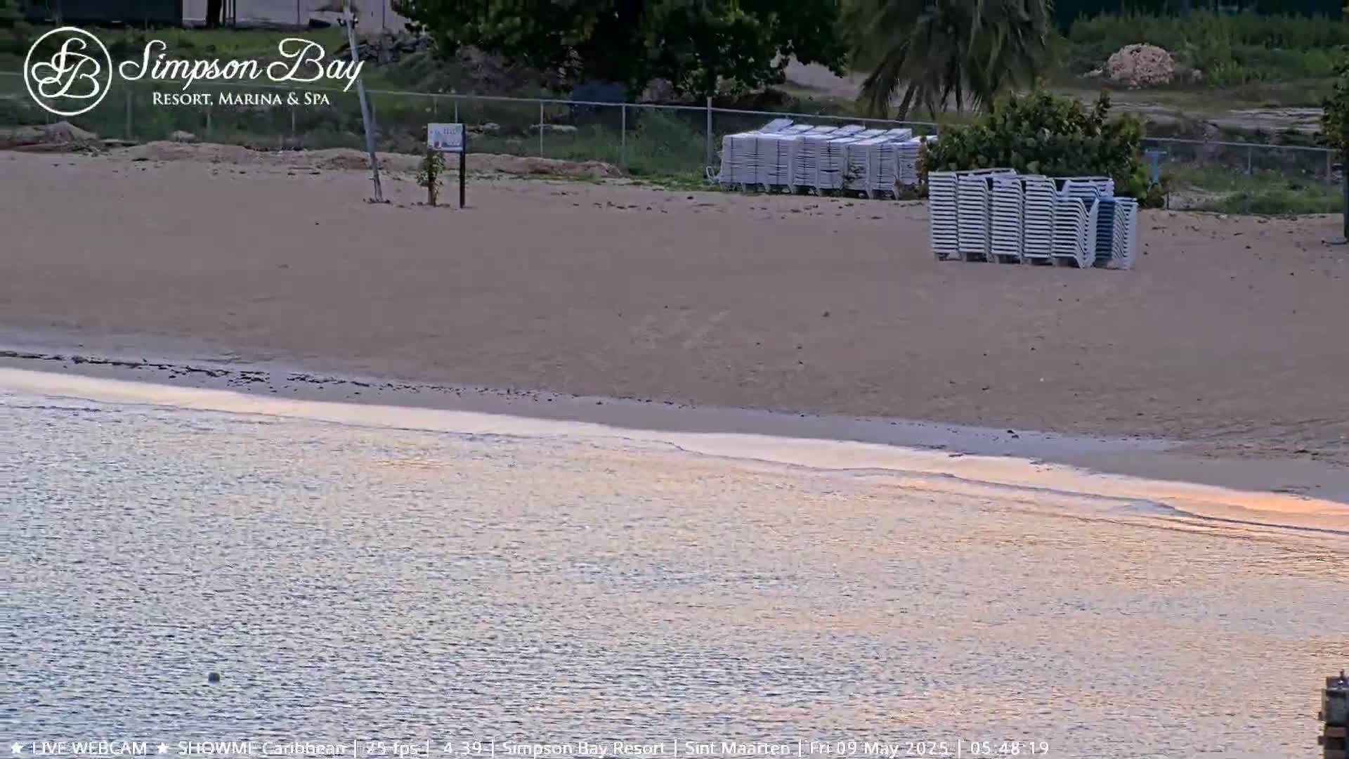 A sandy beach with stacked lounge chairs is next to calm, pale-orange water at sunset.
