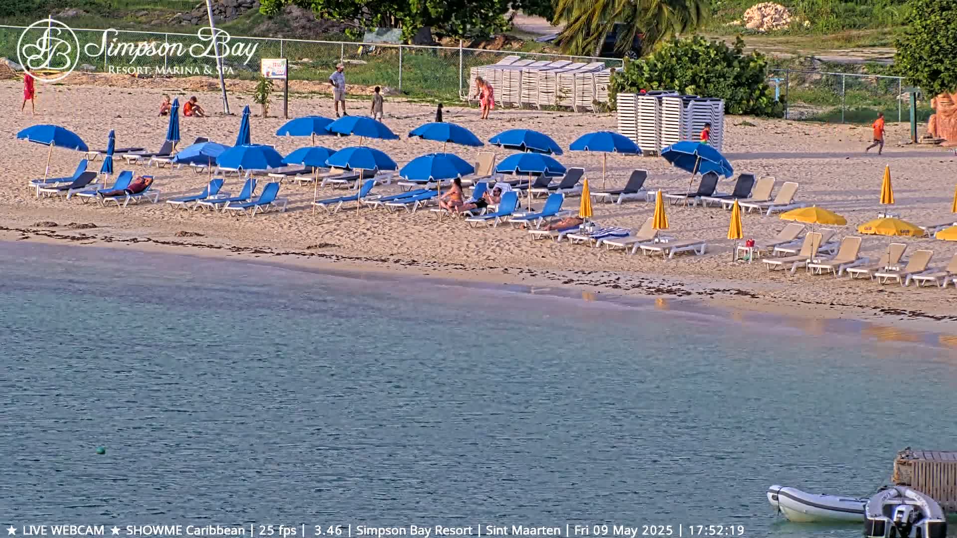 A sandy beach with numerous blue and yellow umbrellas and lounge chairs, a calm body of water, and a few people are visible under a sunny sky.