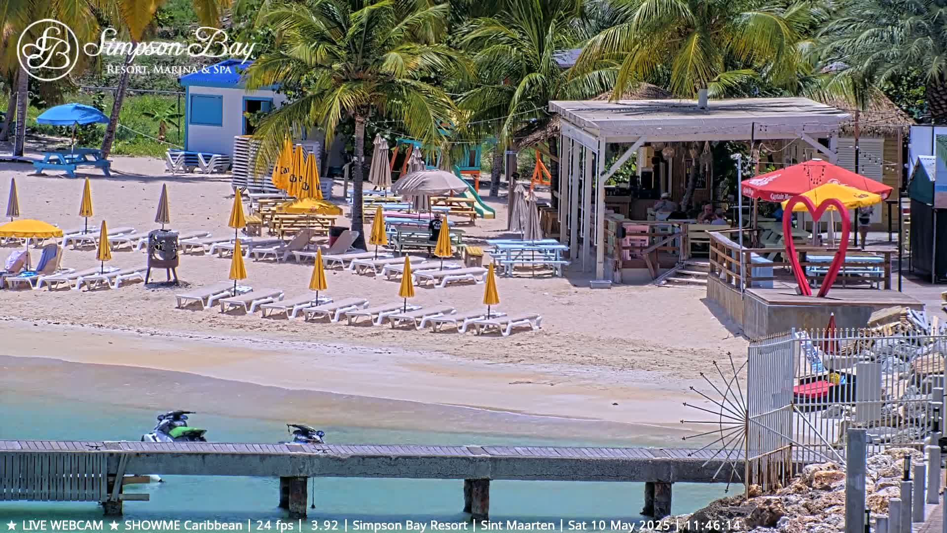A sunny beach scene shows lounge chairs and umbrellas under palm trees, near a beach bar and a small pier with jet skis.