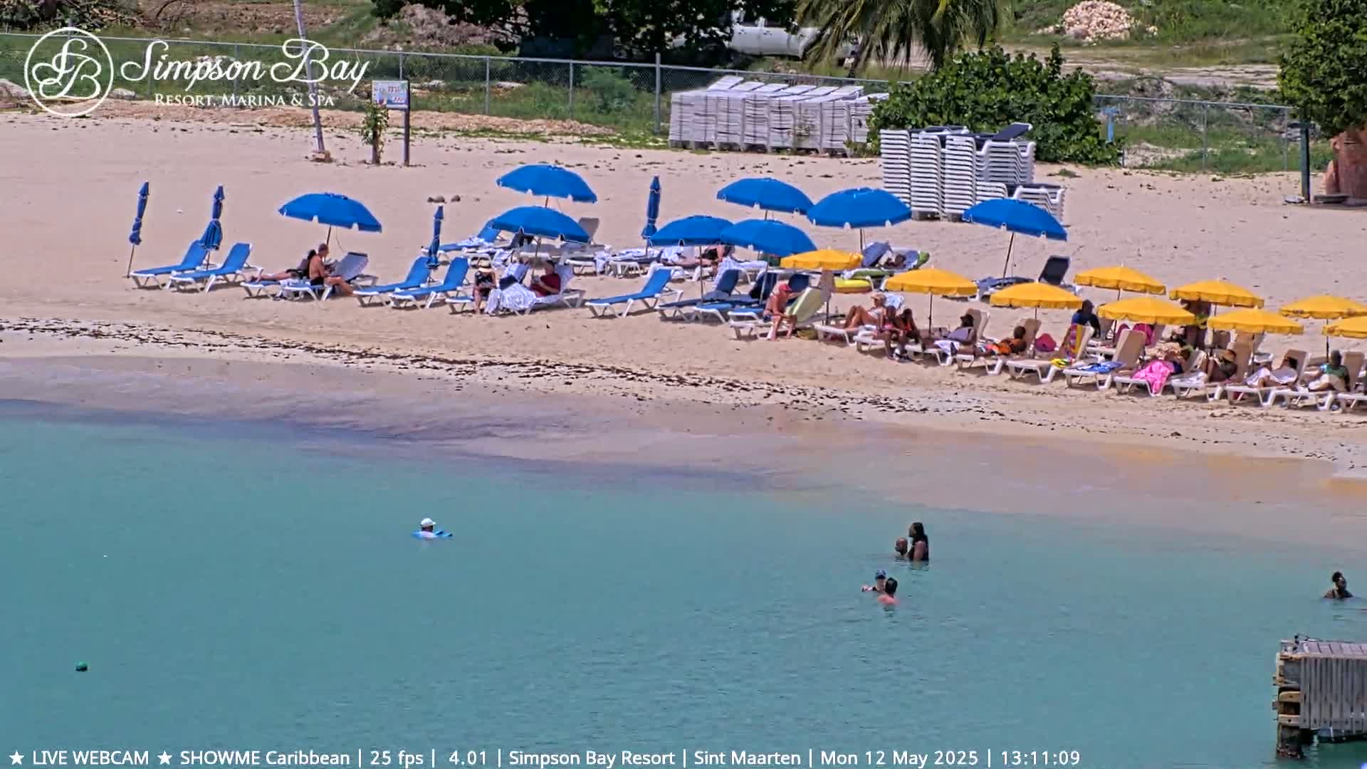 A sandy beach with numerous lounge chairs and umbrellas under a sunny sky, and calm, clear turquoise water with several people swimming.