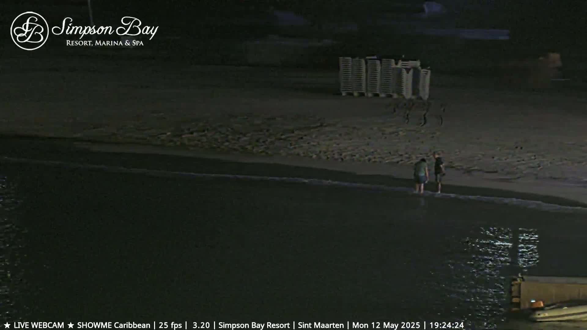 Two people stand at the water's edge on a dark beach at night, near a row of stacked beach chairs.