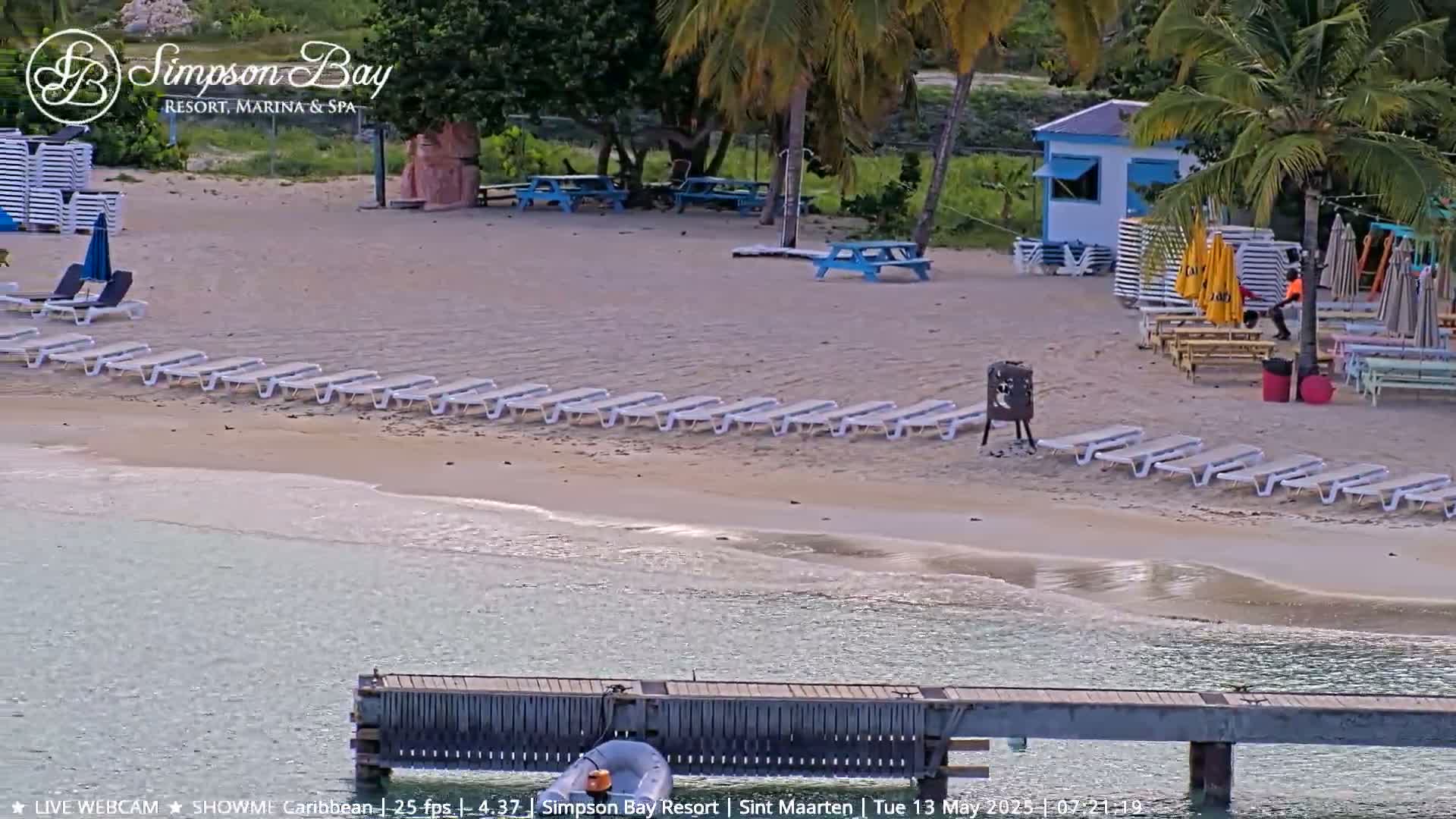 A sandy beach with rows of lounge chairs, picnic tables, and a small building is shown next to calm, shallow water and a wooden dock.