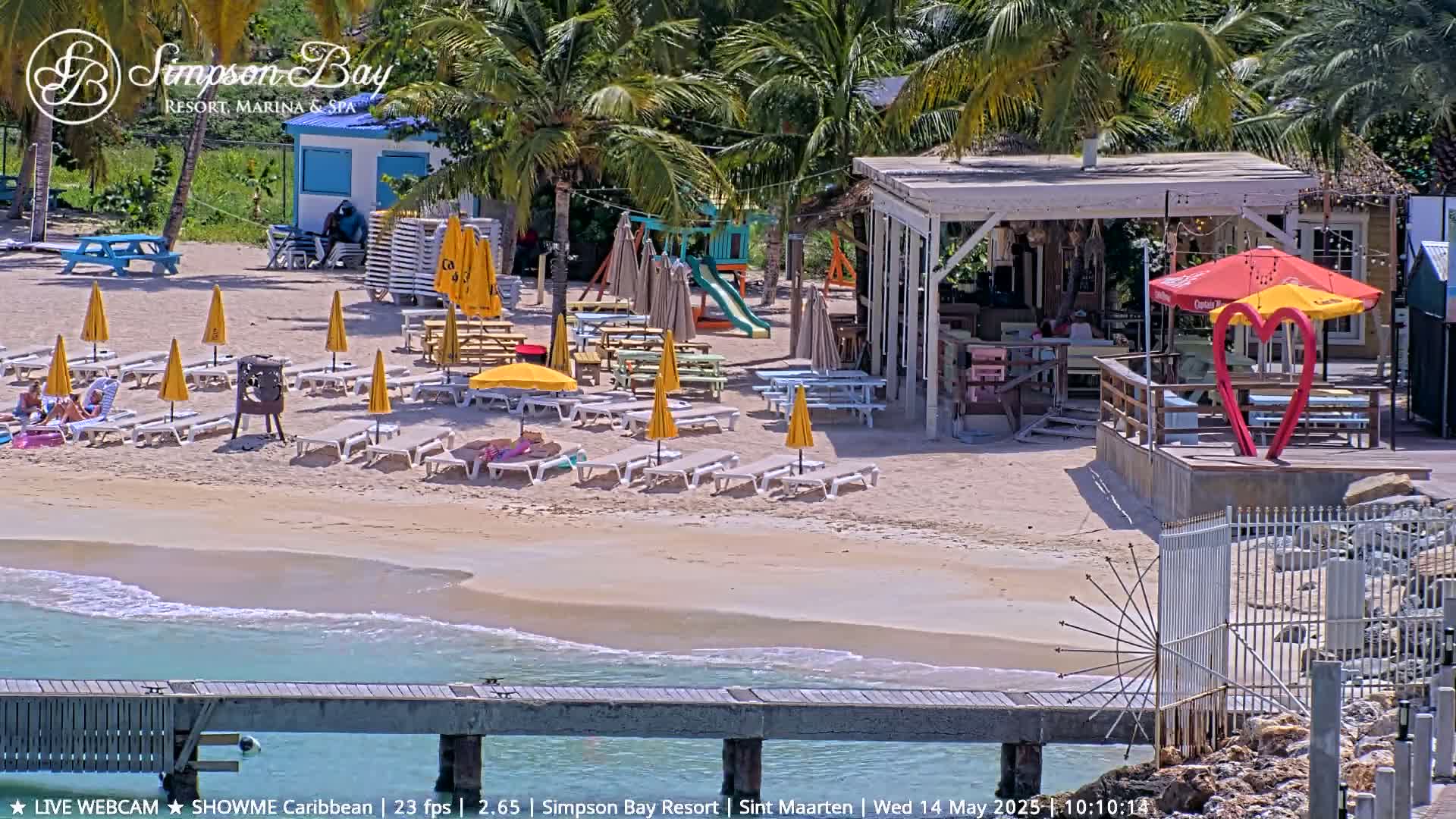 A sunny beach scene shows lounge chairs under yellow umbrellas, a beachside bar, a playground, and calm ocean waves lapping a sandy shore.