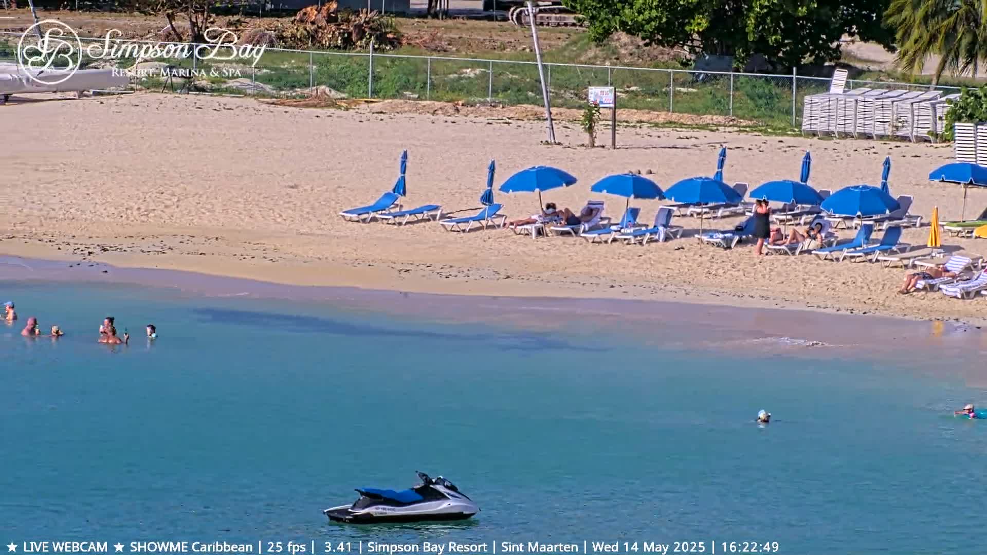 A sandy beach with blue umbrellas and lounge chairs, several people swimming and sunbathing, and a jet ski in calm, clear turquoise water.