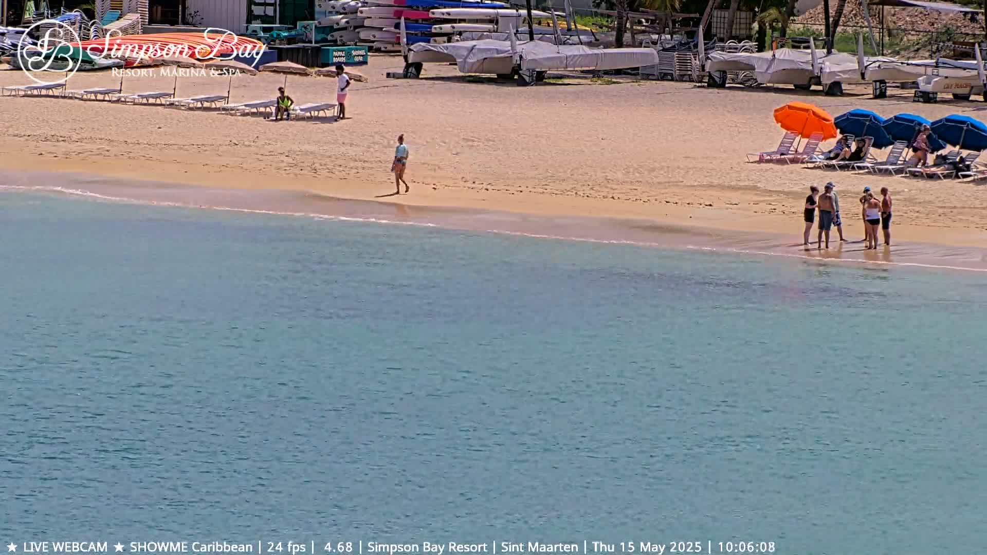 A sandy beach with several people, beach umbrellas, and lounge chairs borders calm, turquoise water; boats are docked in the background.
