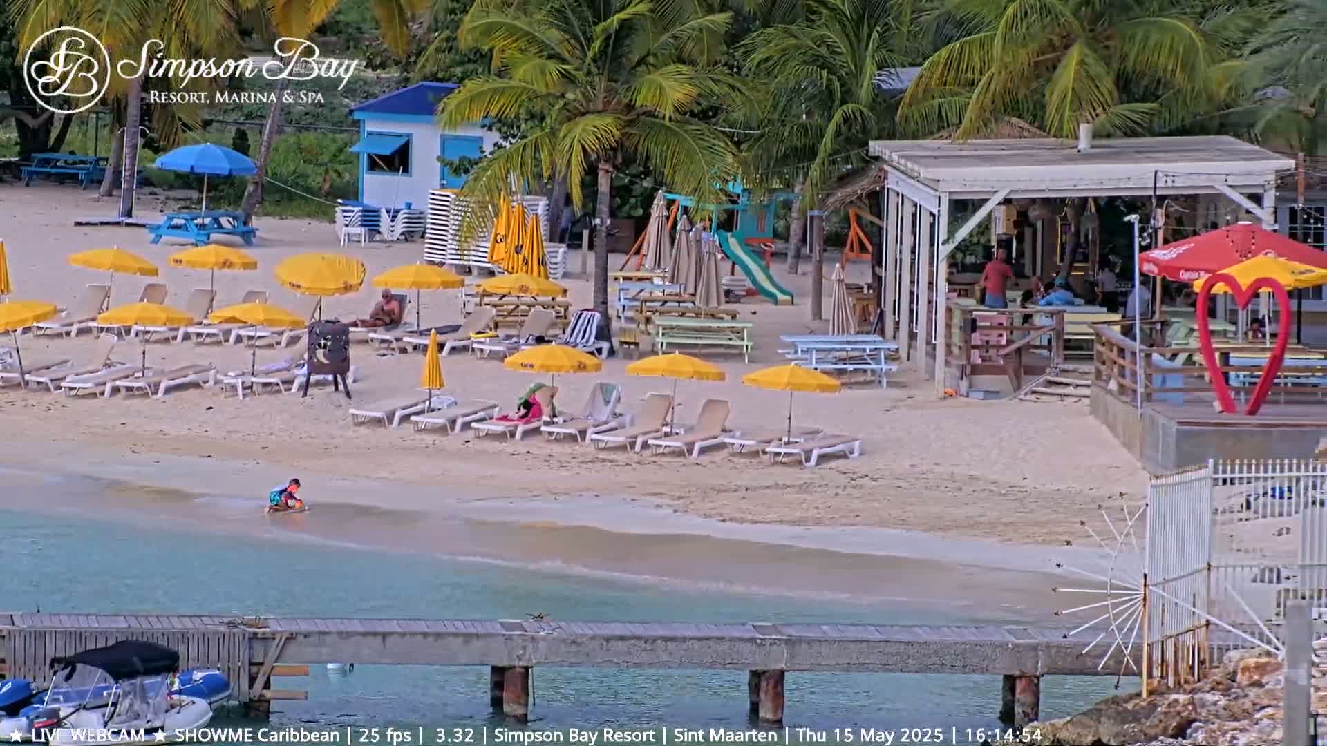 A sandy beach with numerous yellow umbrellas, beach chairs, and a beachfront bar is shown, with calm, shallow, turquoise water lapping at the shore.