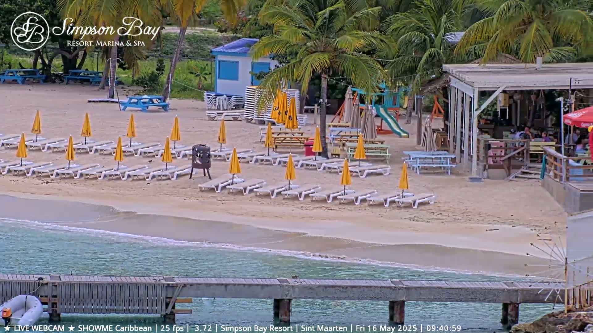A sandy beach with rows of yellow umbrellas and lounge chairs, a beach bar, playground equipment, and a calm ocean with a small pier.