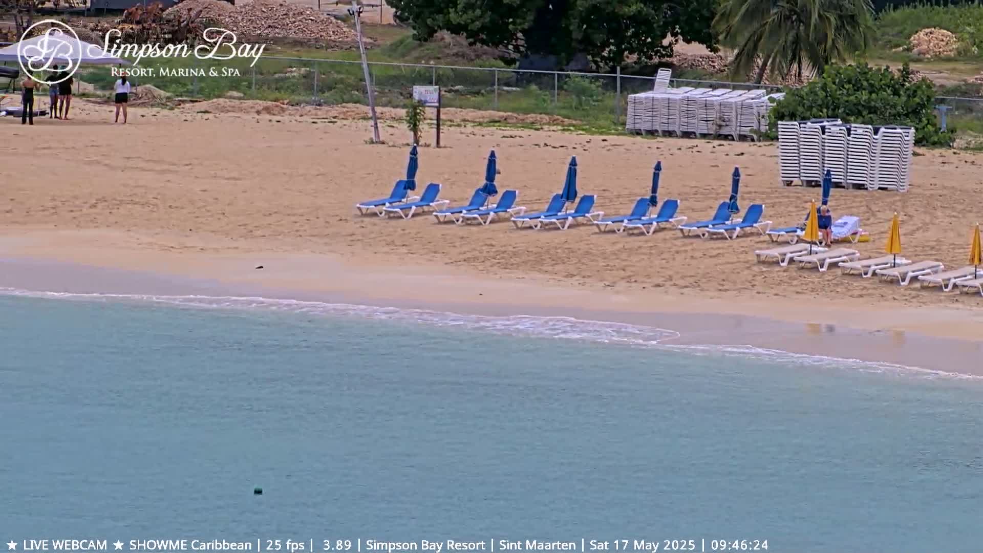 A sandy beach with blue lounge chairs and umbrellas, a few people, and calm, light-blue water.