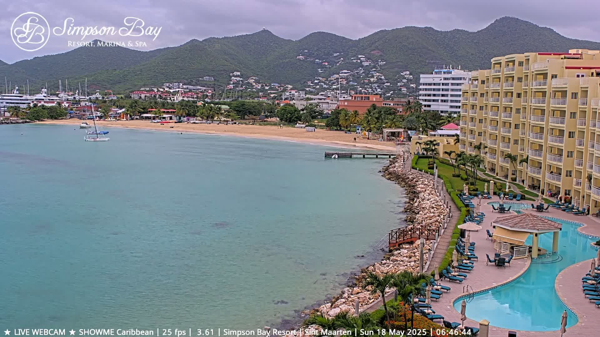 A resort pool and beach are seen next to calm, teal water, with boats in the distance and mountains under a cloudy sky.