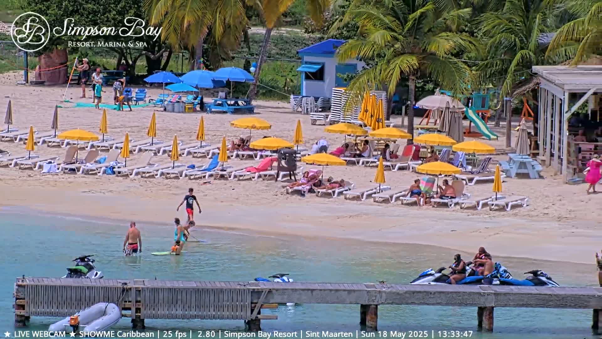 A sunny beach scene shows people relaxing on lounge chairs under yellow umbrellas, others swimming in the calm water, and jet skis docked near a wooden pier.