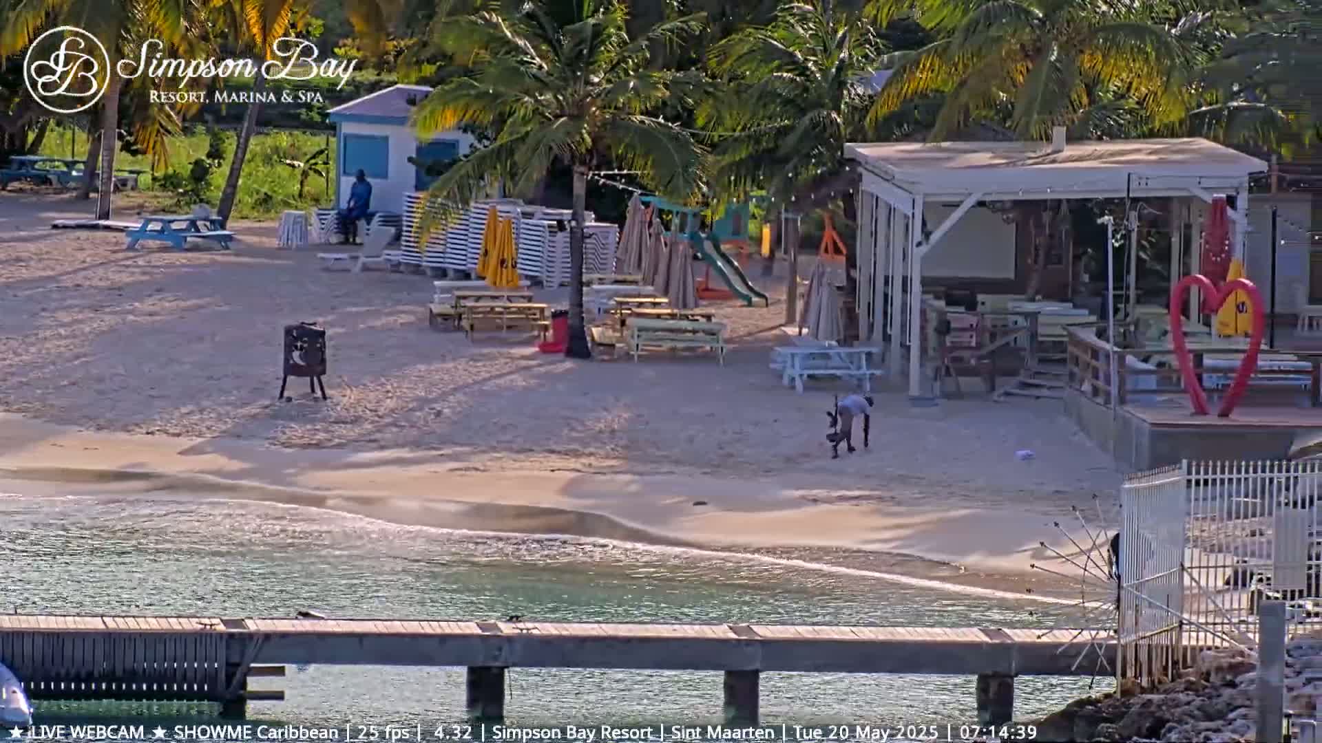 A sandy beach with picnic tables, beach umbrellas, a small playground, and a building with a covered patio is shown on a sunny day next to calm, clear water.