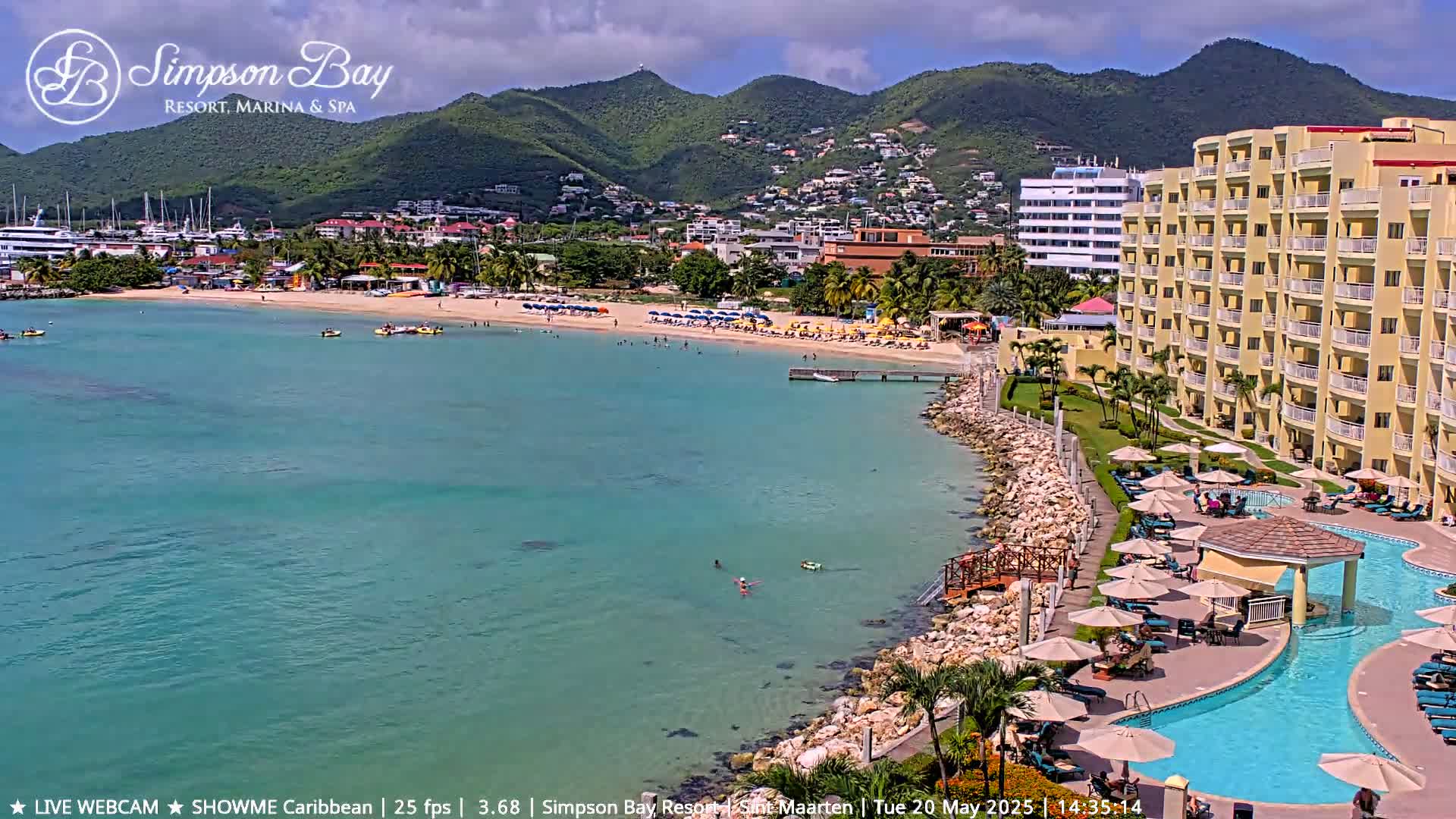 A resort with a swimming pool overlooks a calm, turquoise bay with a sandy beach and a town nestled in green hills under a partly cloudy sky.