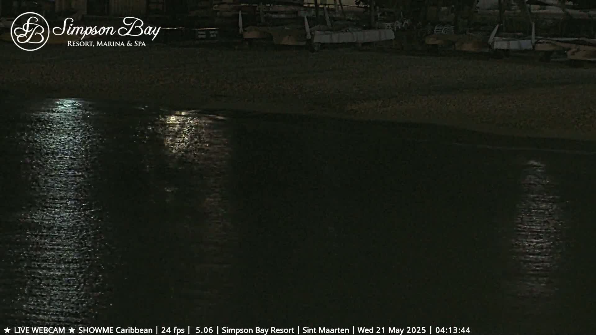 A dark, moonlit beach at night shows calm water reflecting the moon's light, with several dark objects resembling boats or beach equipment lined up on the sand.