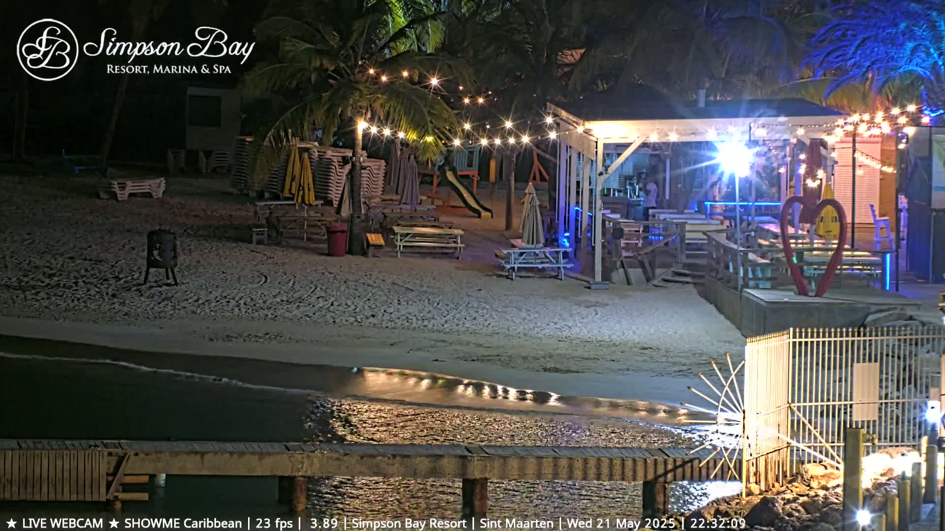 A nighttime beach scene shows a bar and restaurant with string lights, picnic tables, and lounge chairs, next to a calm body of water under a clear sky.