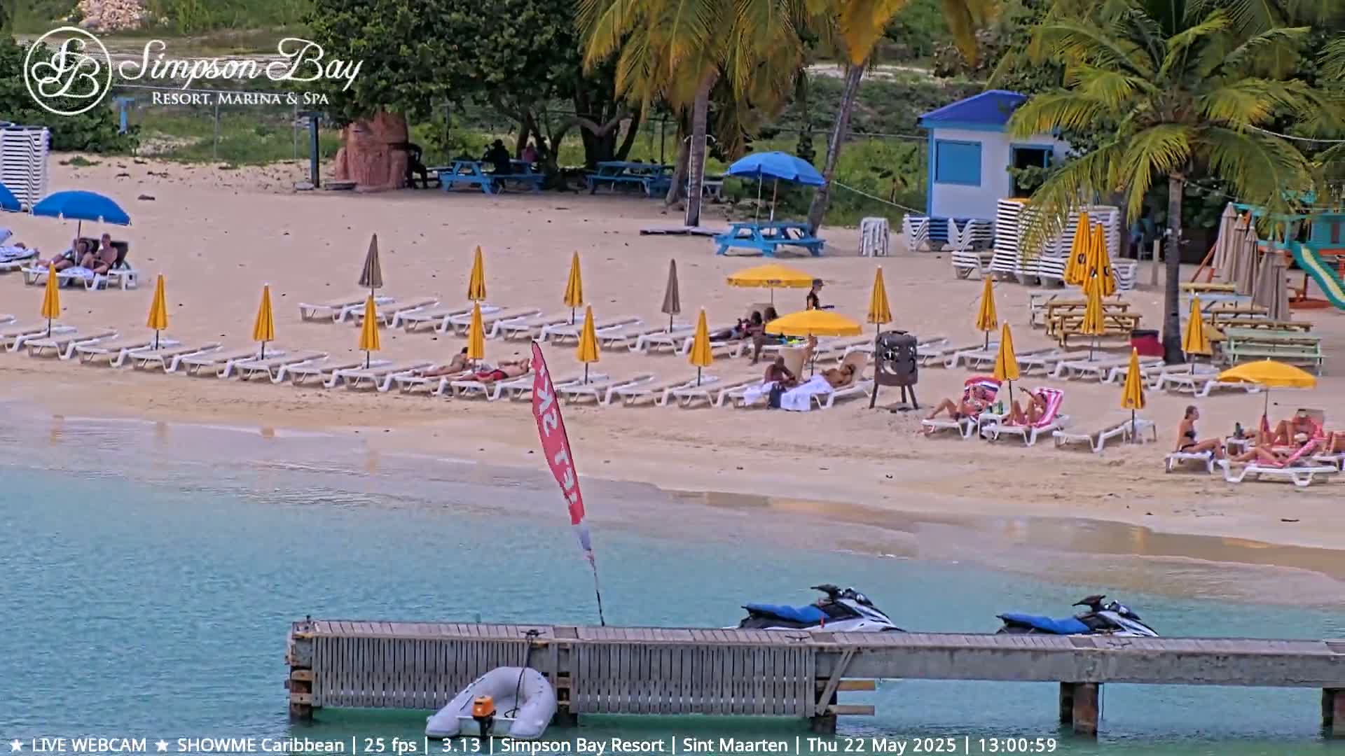 A sandy beach with numerous lounge chairs and umbrellas under a sunny sky, next to calm turquoise water with jet skis docked at a pier.