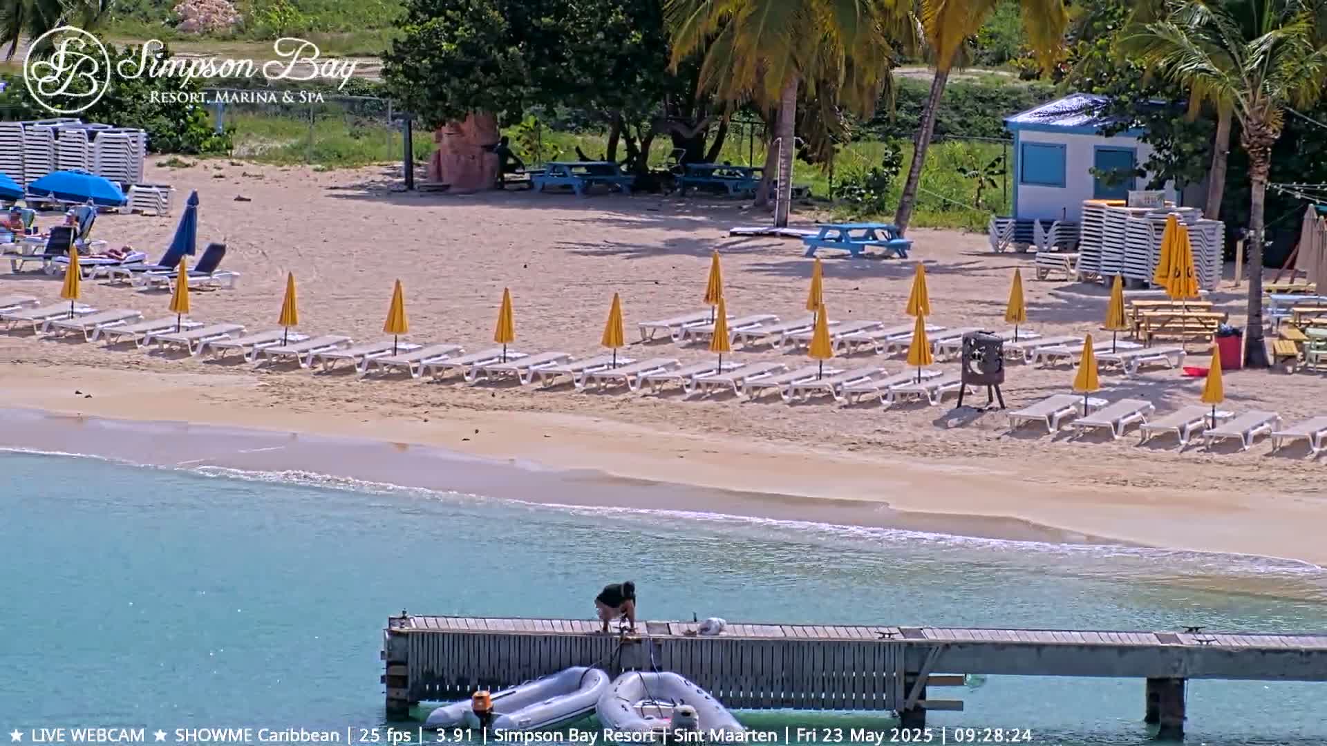 A sandy beach with rows of lounge chairs and yellow umbrellas, a small building, and a person on a dock next to calm turquoise water.