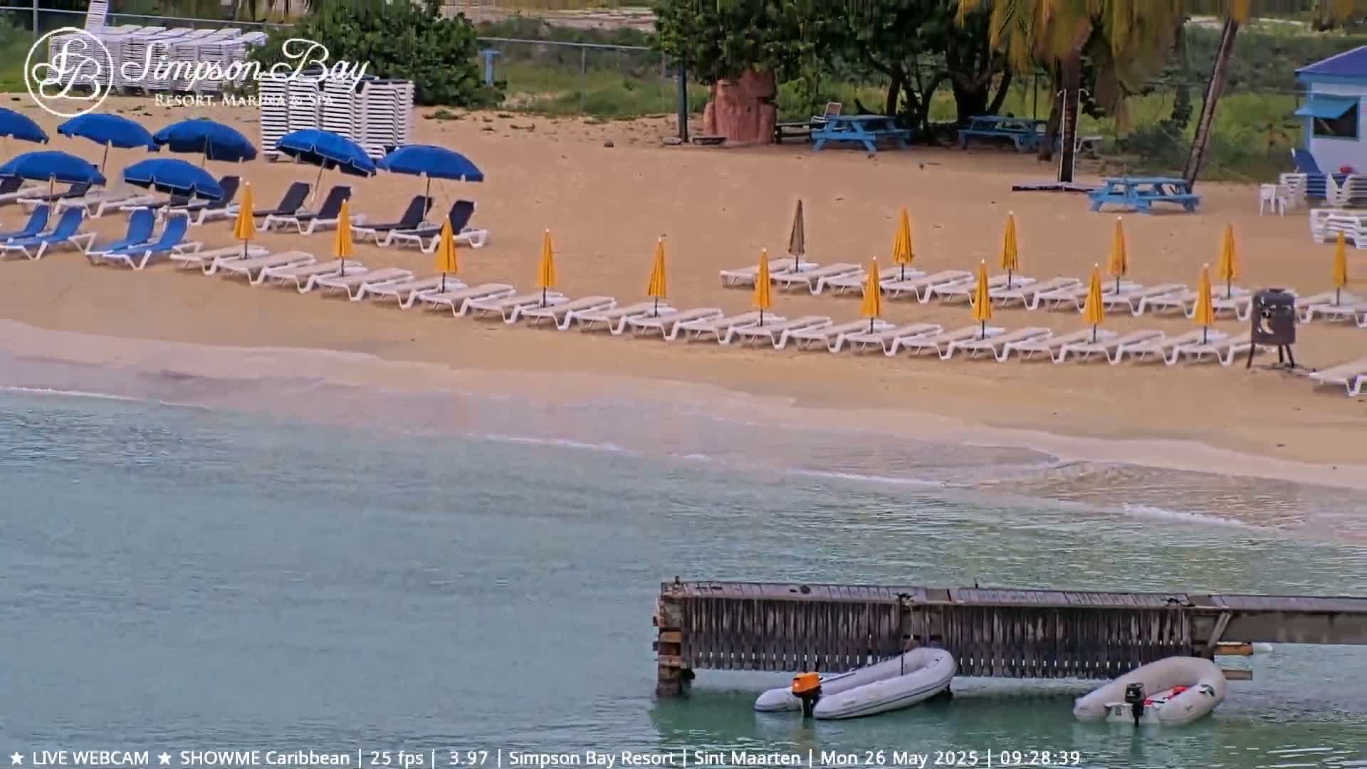 A sandy beach with rows of lounge chairs and umbrellas under a partly cloudy sky, next to calm turquoise water with a small wooden dock and two small boats moored to it.