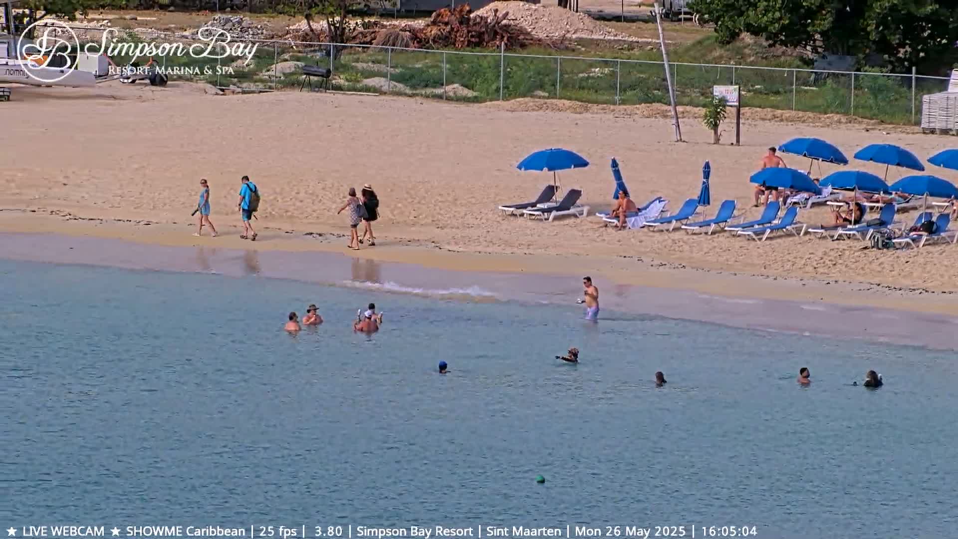 A sandy beach with calm, clear water has people walking, lounging in beach chairs under umbrellas, and swimming.