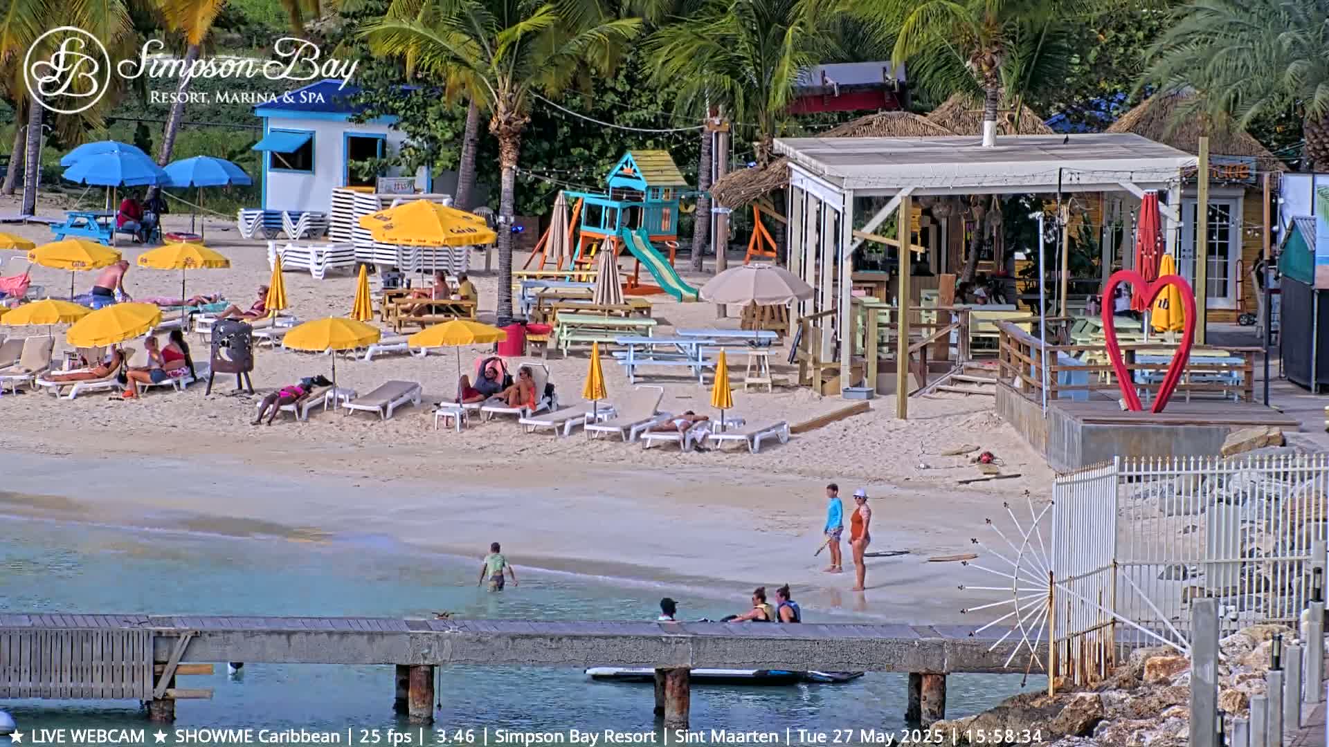 A sunny beach scene shows people relaxing under yellow umbrellas, near a beach bar, playground, and a dock extending into calm, clear water.