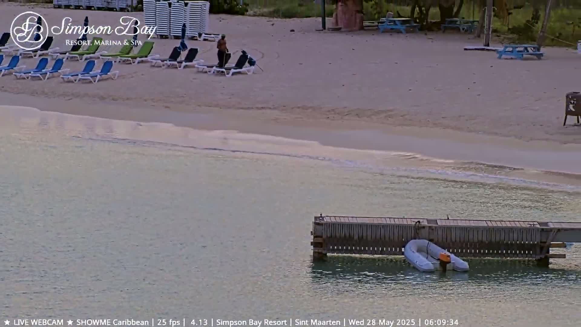 A sandy beach with lounge chairs and a person sweeping, next to calm, shallow water with a small inflatable boat docked at a pier.