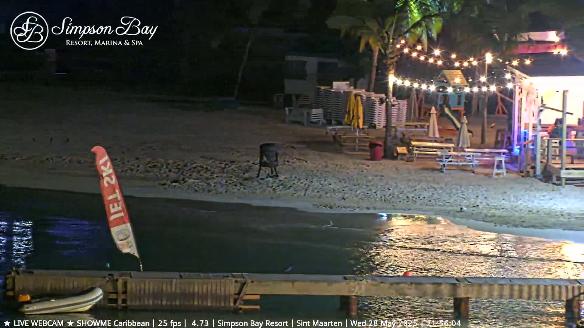A nighttime beach scene shows a calm body of water lapping at a sandy shore, where picnic tables and a small building with string lights are situated near a dock with a small boat.