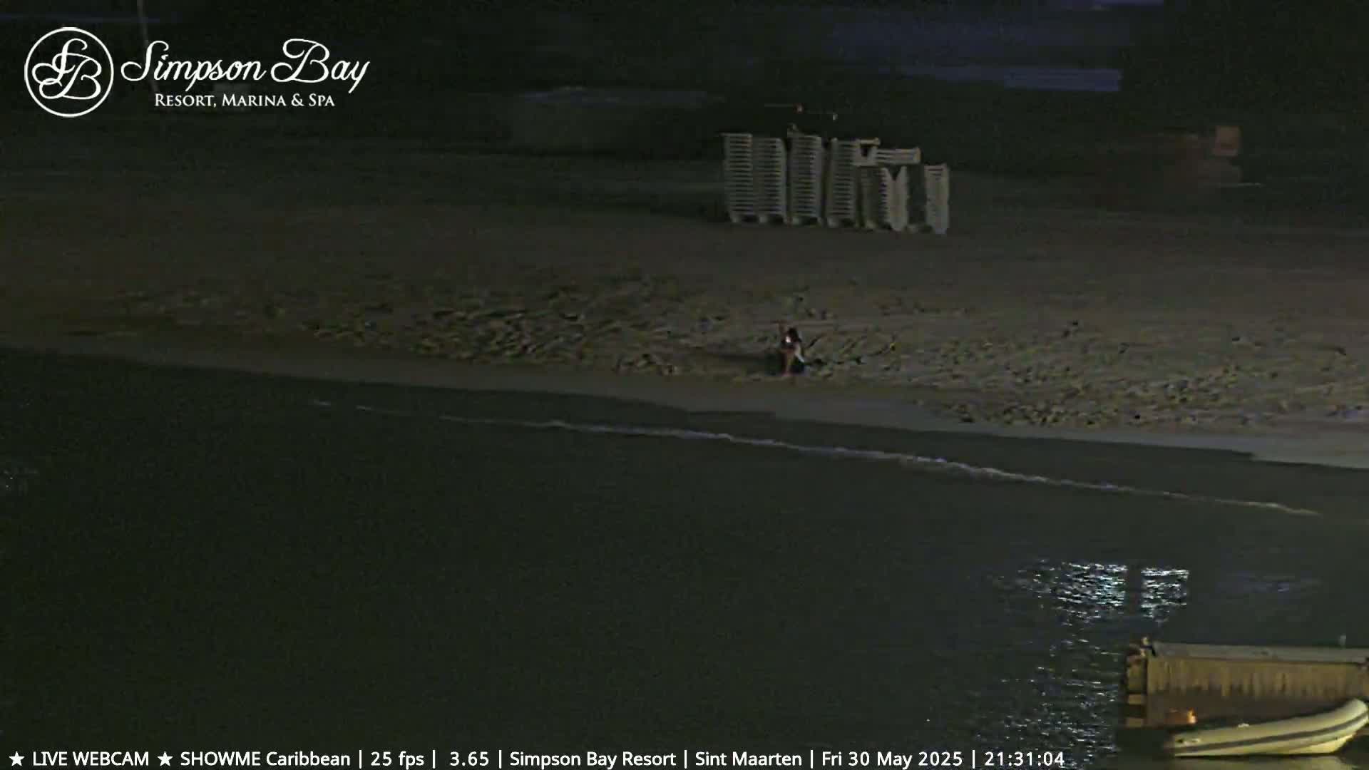 A mostly dark sandy beach at night with calm water, a few beach chairs stacked together, and a single person sitting on the sand.